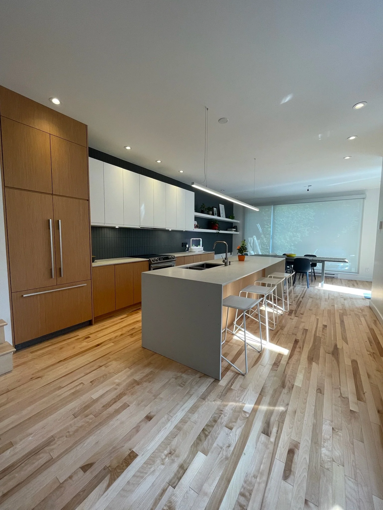 Modern kitchen with wooden cabinetry, white upper cabinets, a kitchen island with a sink, bar stools, a dining table with chairs, and large windows letting in natural light.