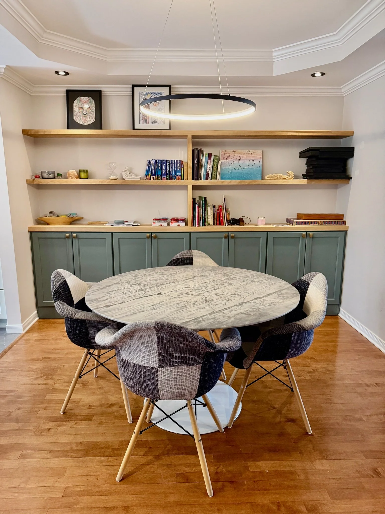 A dining room with a round marble table surrounded by six modern, upholstered chairs with wooden legs. Behind the table, there is a light green cabinet with storage doors, and above it are three wooden shelves holding books, artwork, and decorative i