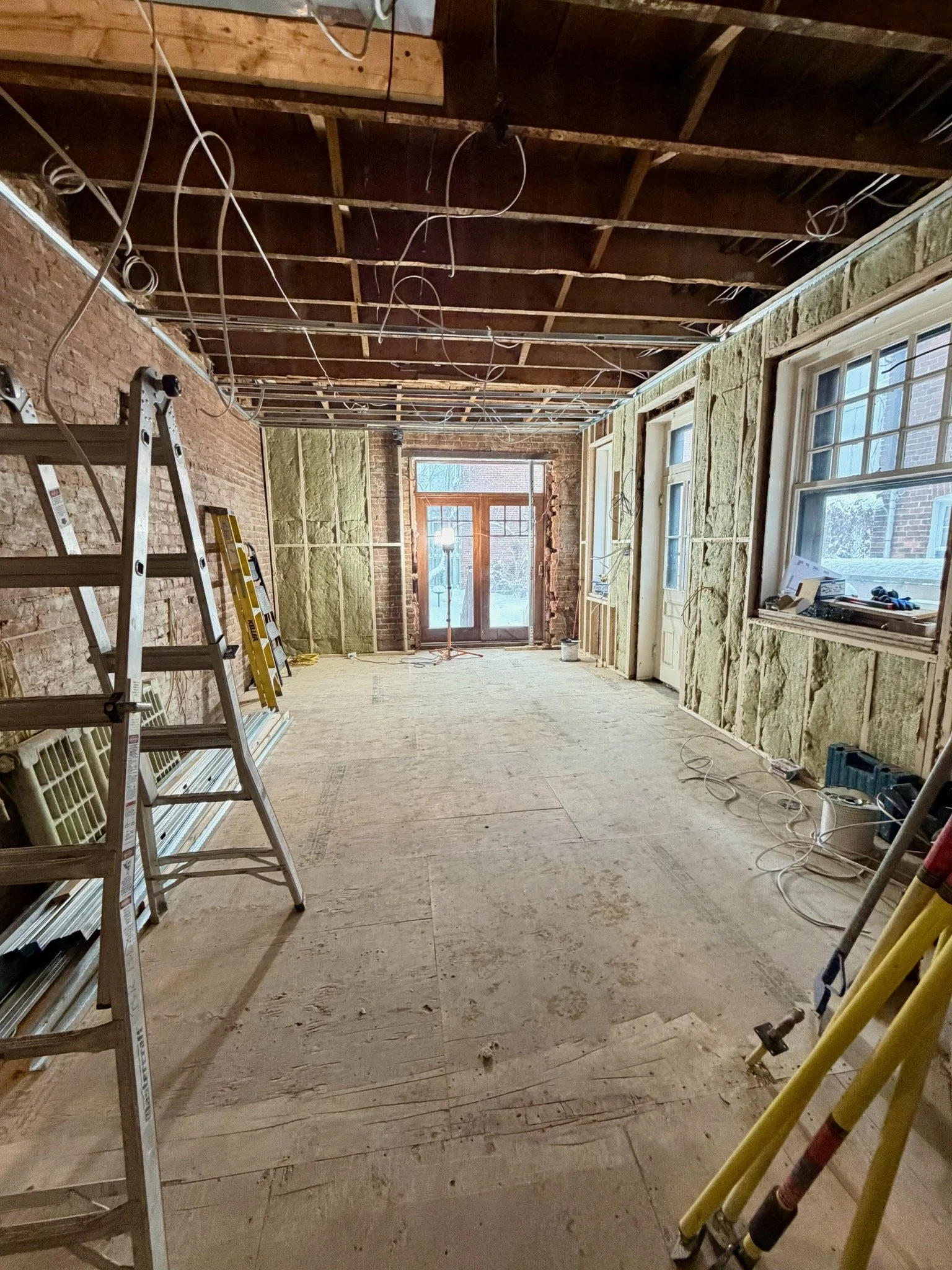 Interior of a room under renovation with exposed brick and insulation on the walls, unfinished ceiling with electrical wiring, windows, and construction ladders.