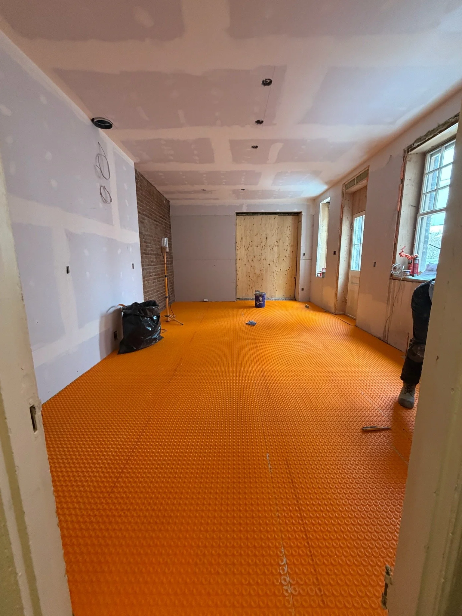 Interior of a room under renovation with an orange rubber floor, unfinished walls, and a ceiling with visible drywall patches. There are windows on the right side and construction tools and materials scattered around.