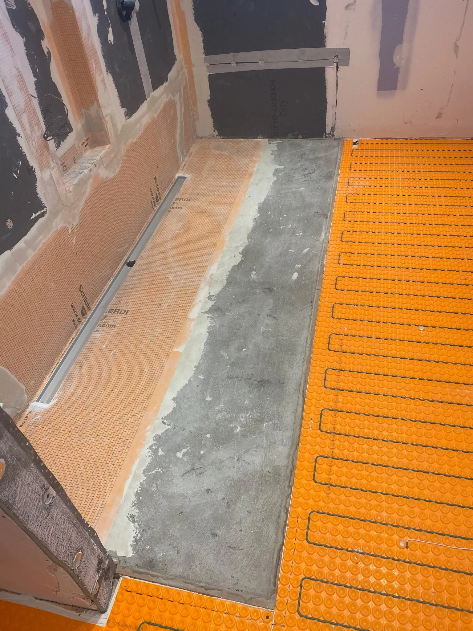 This image shows a partially completed bathroom or similar space under construction. The floor has orange safety tiles with black cables arranged on top. The walls are unfinished, with some insulation and subwall visible. There is a black box or pane