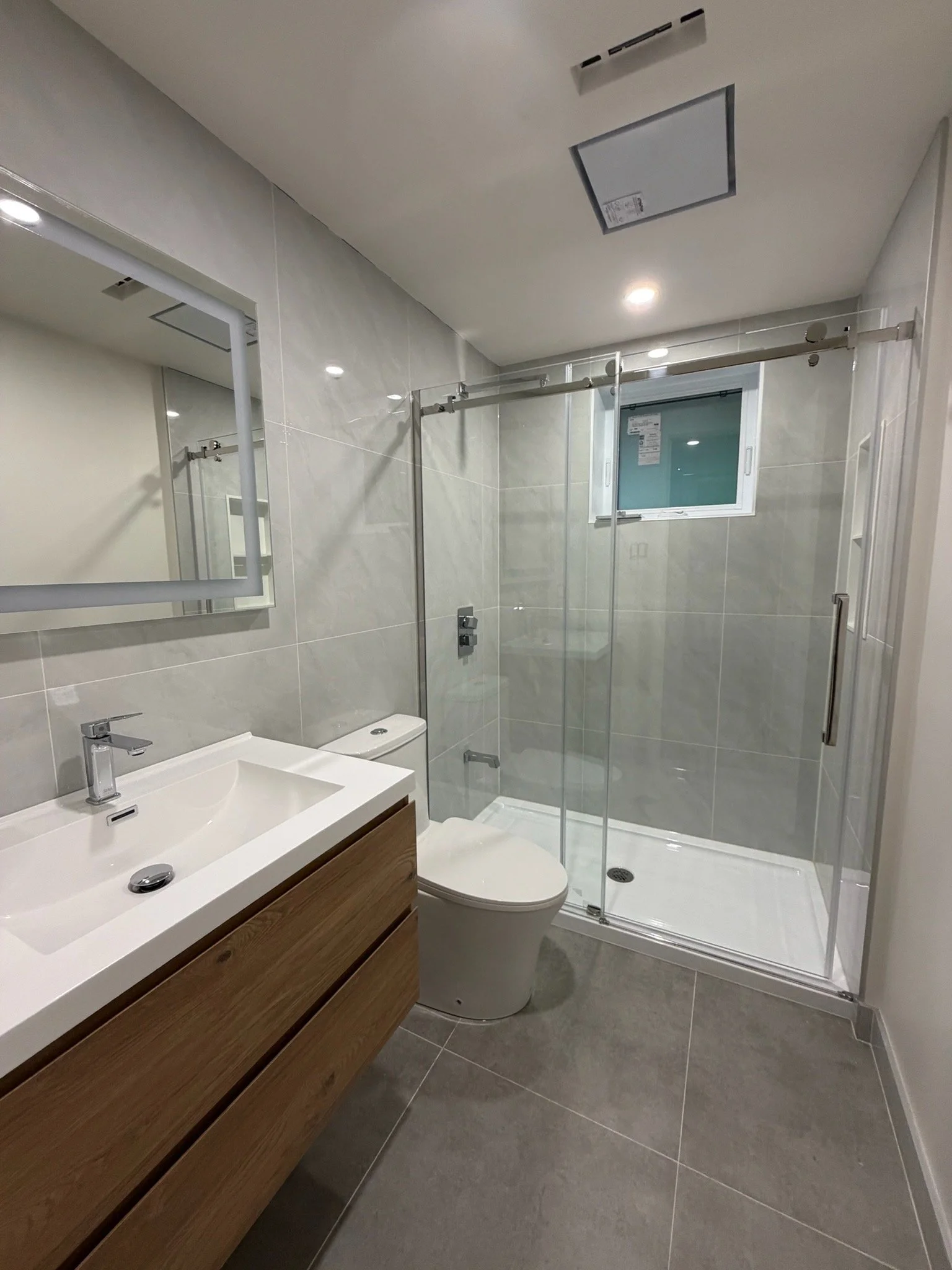 Modern bathroom with a white sink, wooden vanity, toilet, shower with glass door, small frosted window, mirror, and ceiling lights.