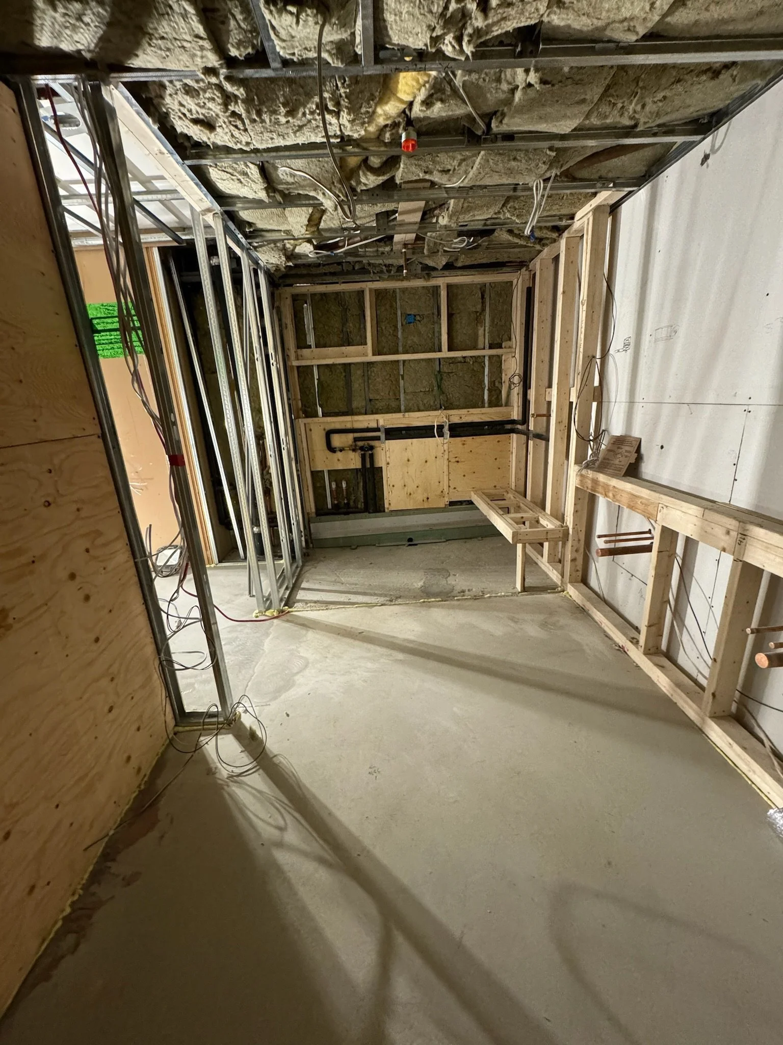 Interior of a room under construction with wooden framing and exposed insulation, wires, and metal pipes visible on the ceiling.