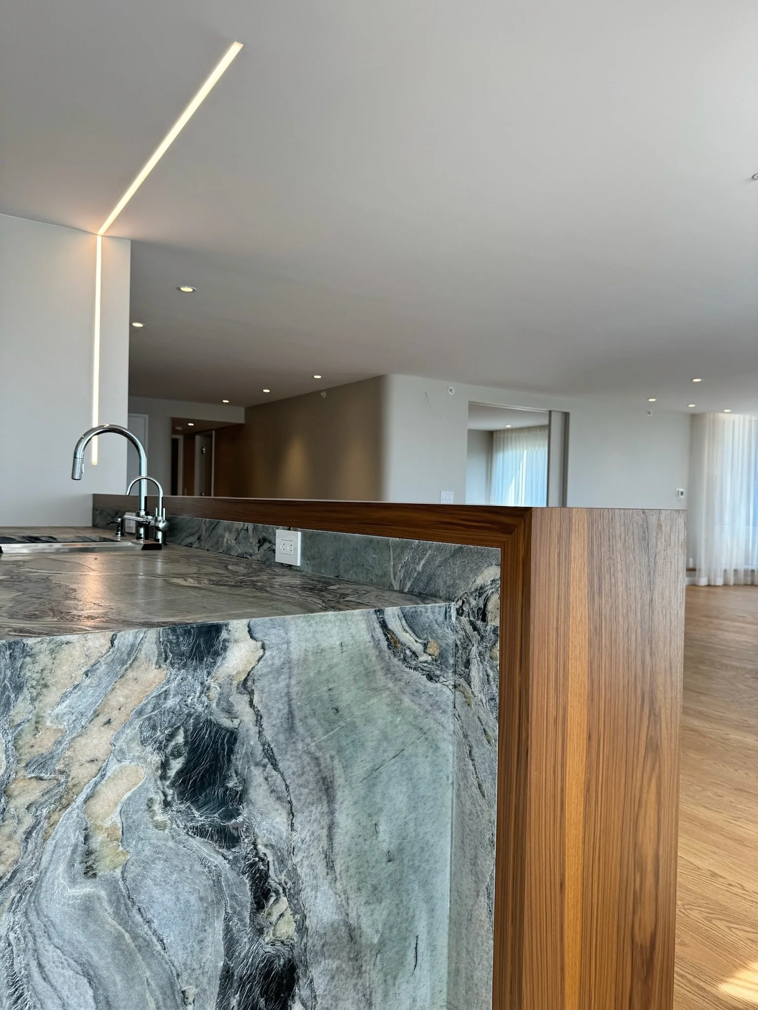Modern kitchen with marble island, stainless steel sink, wooden trim, and open living space in the background with large windows and curtains.