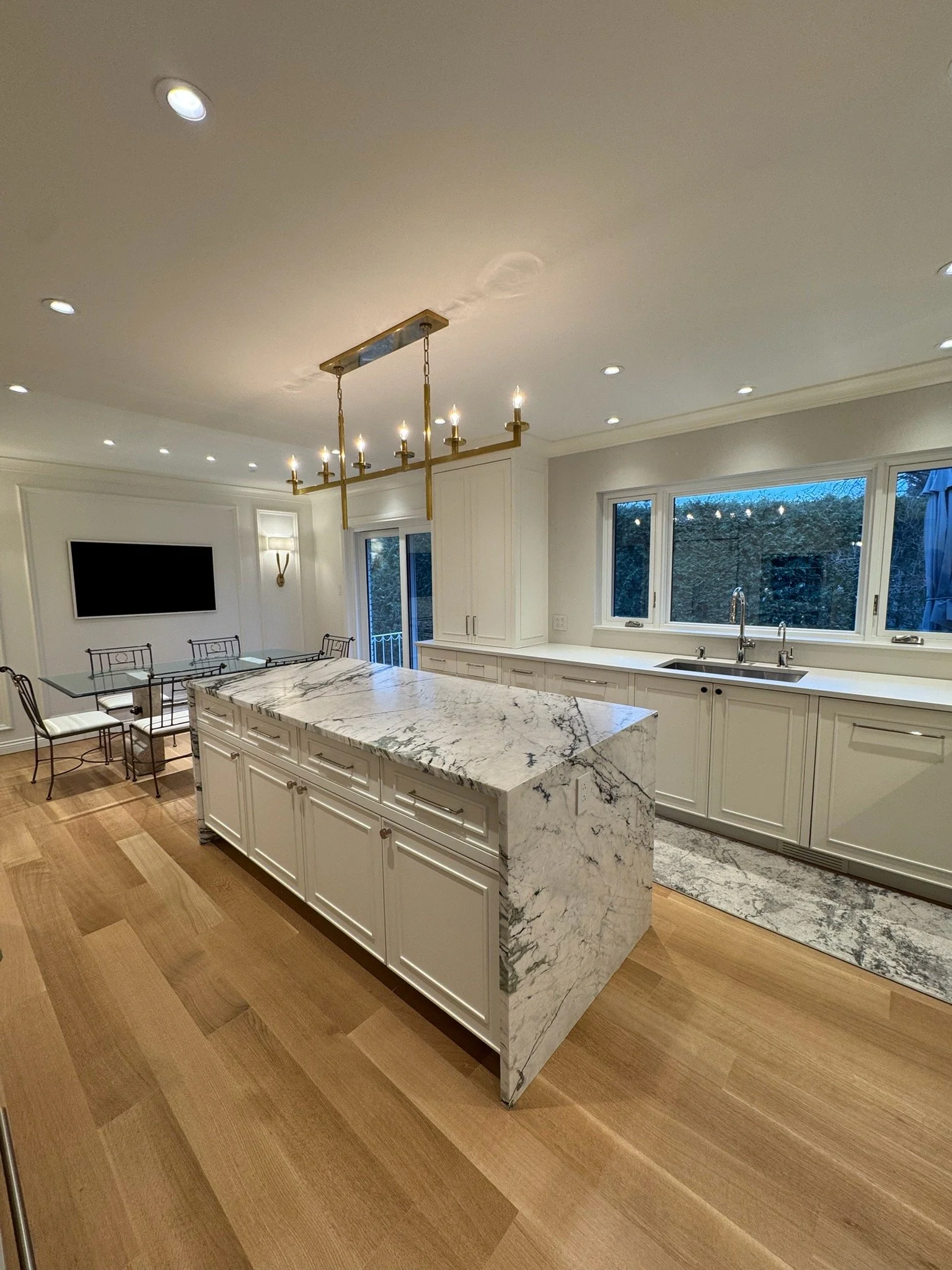 Modern kitchen with white cabinets, marble island, large window, pendant chandelier, hardwood flooring, and a wall-mounted TV.