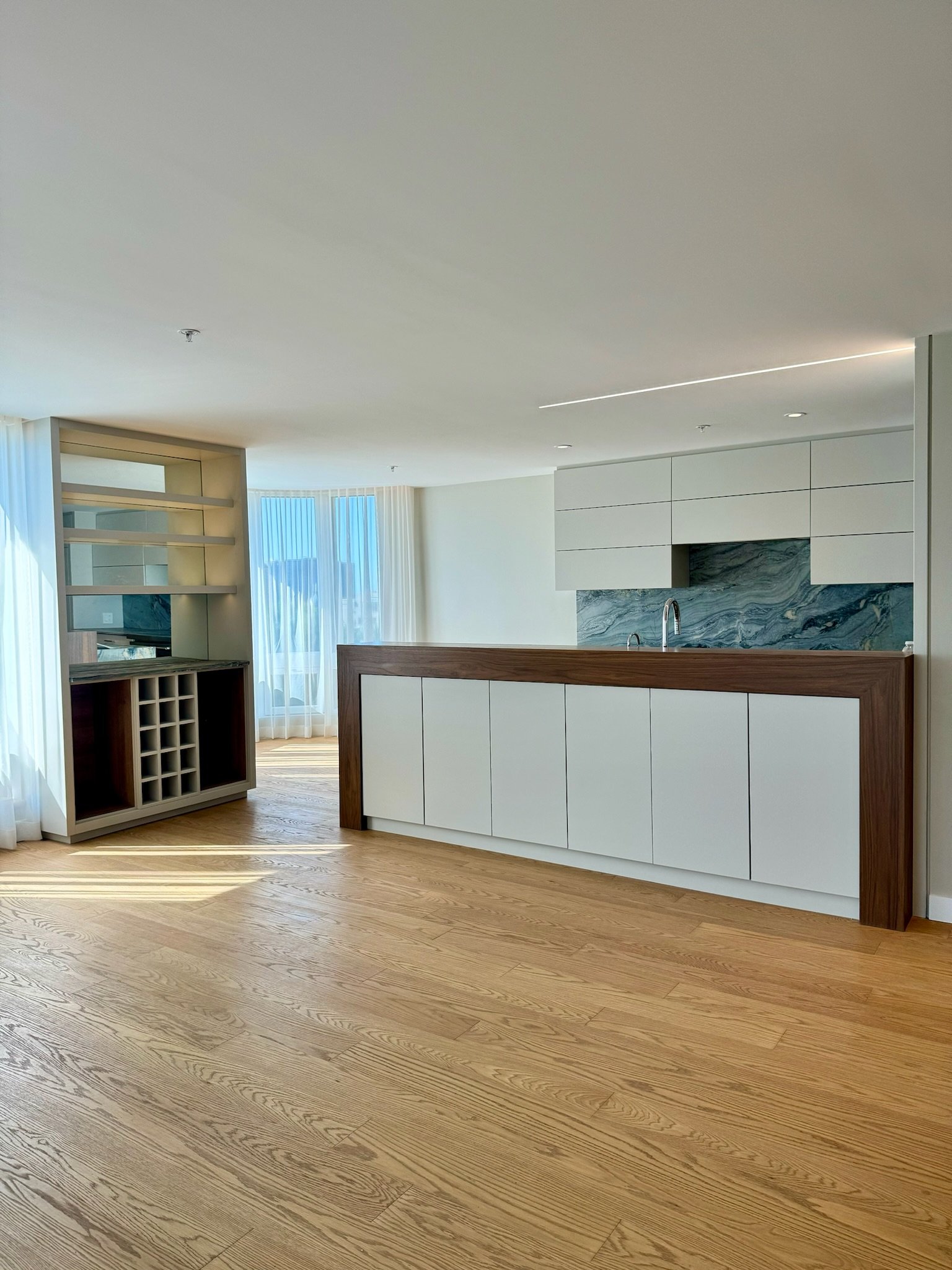 Modern open-concept living space with wooden flooring, large windows with sheer curtains, and minimalist kitchen cabinetry with a marble backsplash.