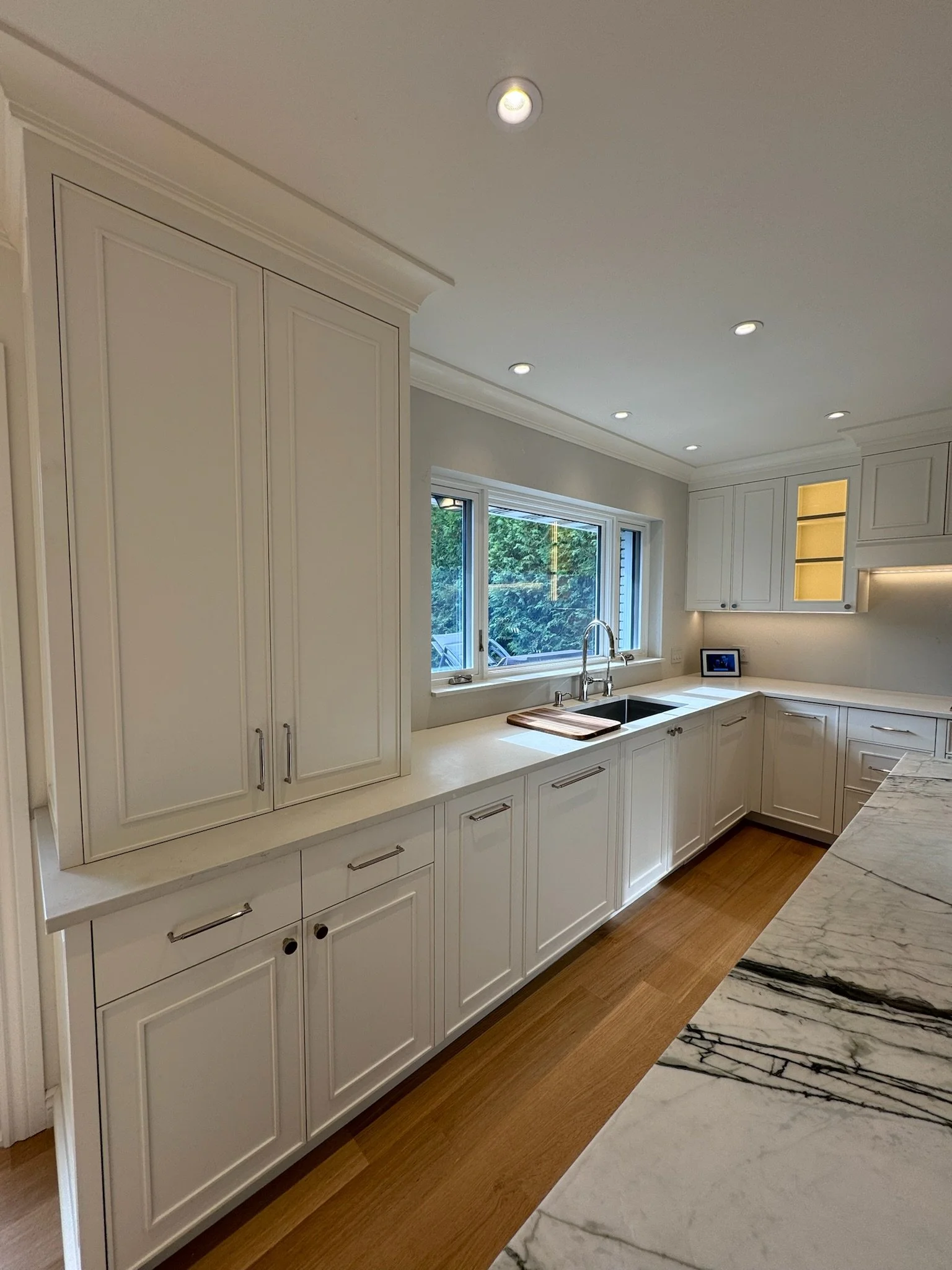 A modern kitchen with white cabinets, a marble countertop, a sink underneath a window, and recessed ceiling lights.