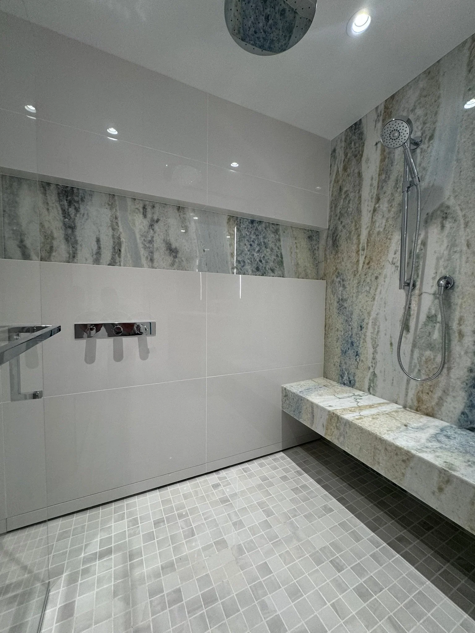 Modern bathroom shower area with marble walls, a handheld shower head, a glass partition, and a tiled floor.