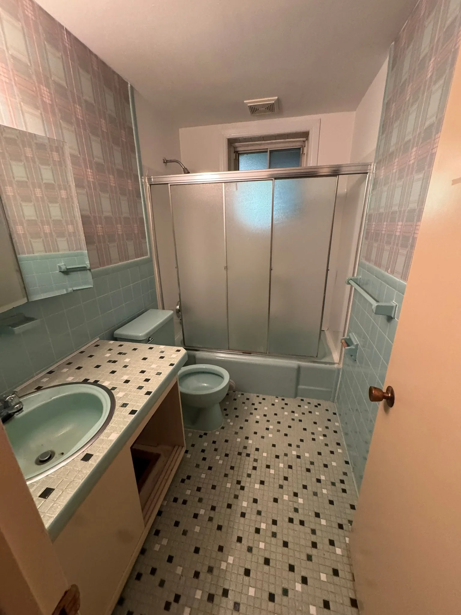 Bathroom with green toilet and sink, tiled floor and half tiled walls, shower with frosted glass door, window above shower, and cabinet under sink.