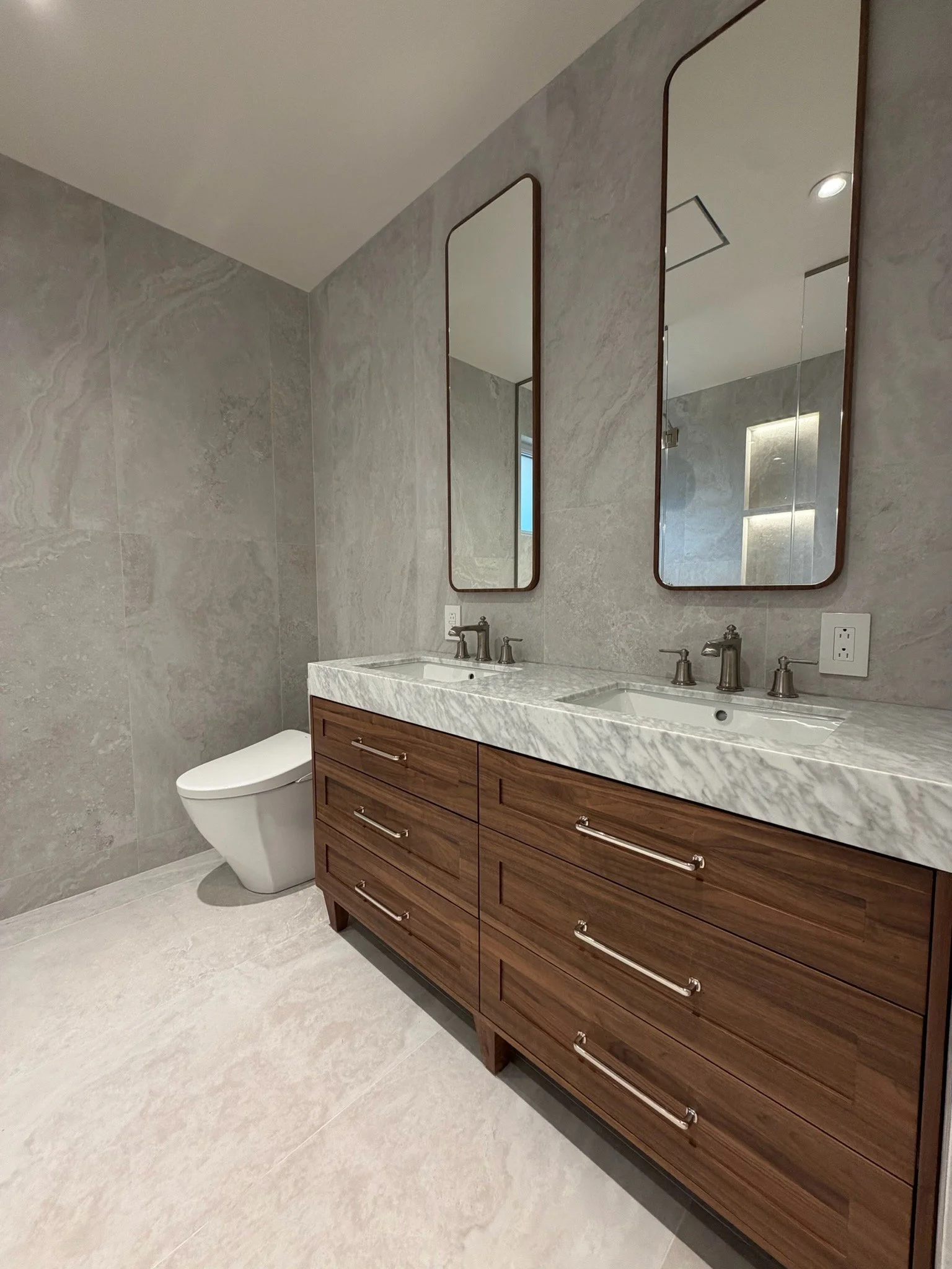 Modern bathroom with a double vanity featuring a marble countertop, two rectangular mirrors with rounded edges, and a toilet. The wall and floor are tiled in light gray.