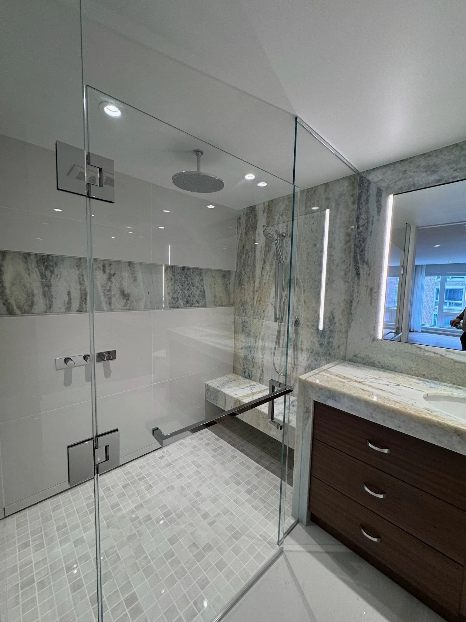 Modern bathroom with a glass-enclosed shower, marble walls, a showerhead, and a vanity with a mirror and drawers.