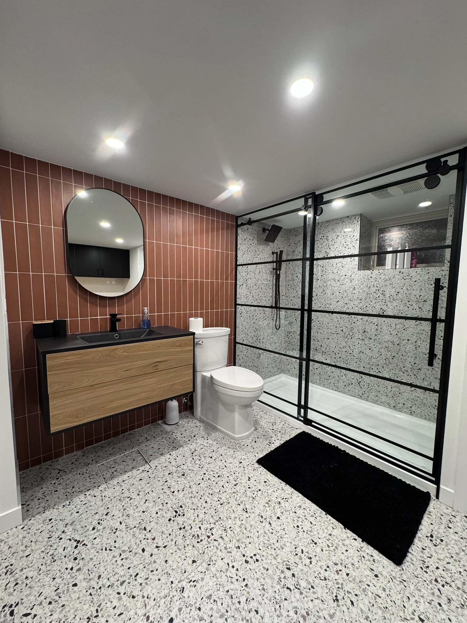 Modern bathroom with terrazzo flooring, brown tiled accent wall, wall-mounted wooden vanity with black countertop, oval mirror, white toilet, walk-in shower with a black metal frame and pebble-patterned wall tiles, black bath mat, and recessed ceilin