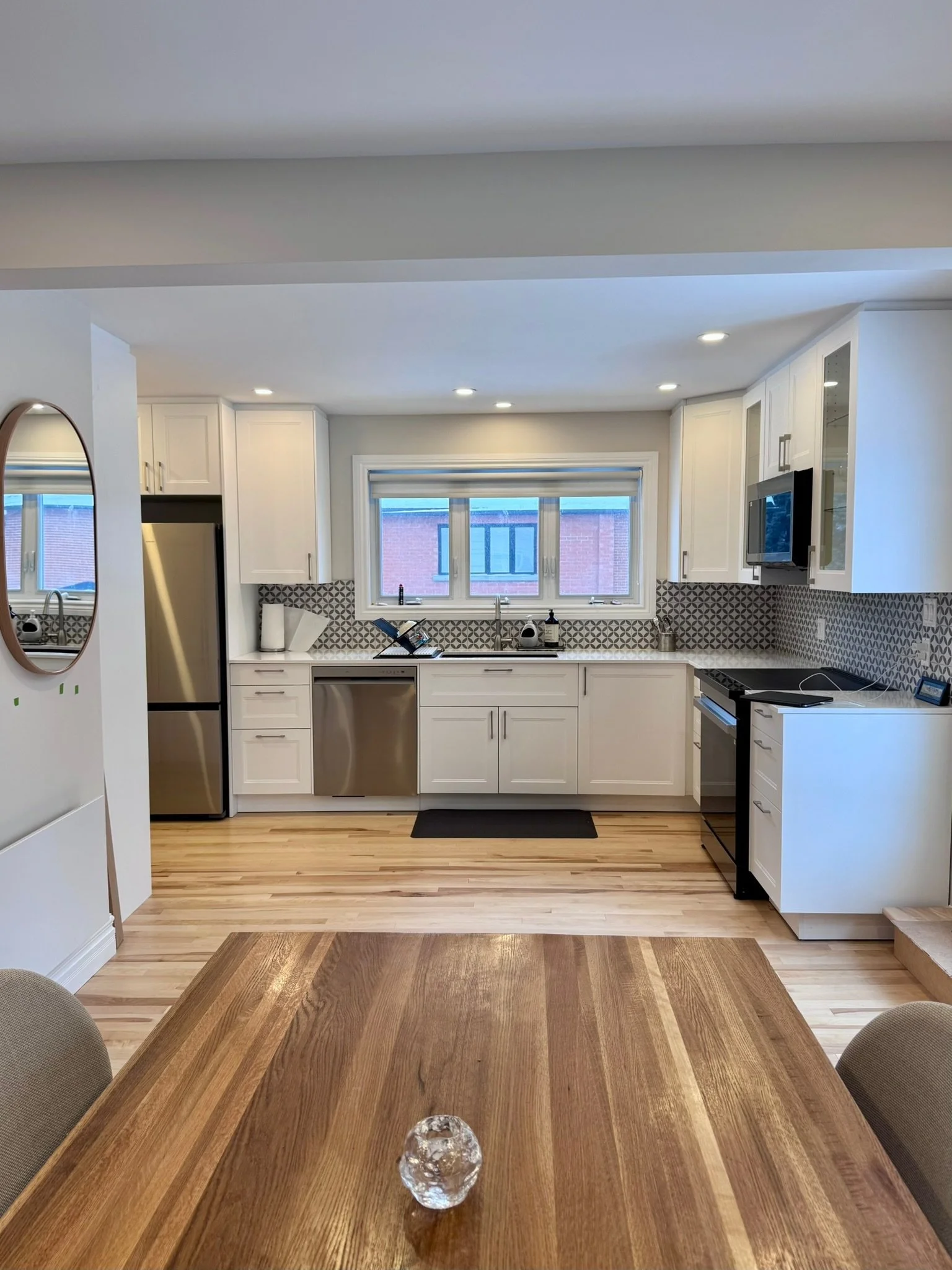 Modern kitchen with white cabinets, black and white geometric backsplash, stainless steel appliances, and a window above the sink. A wooden dining table with a glass decorative piece in the center is in the foreground.