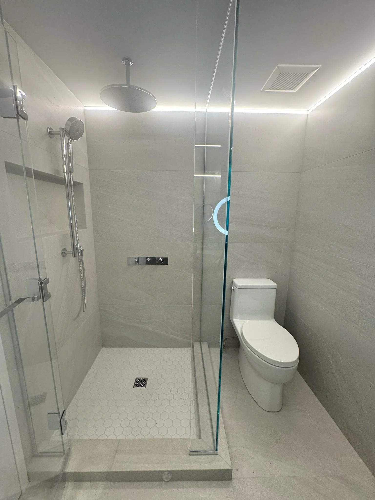 A modern bathroom featuring a glass shower enclosure, rainfall showerhead, handheld shower, a white toilet, beige tiled walls and floor, a drain on the shower floor, a ventilation fan, and minimalistic design.