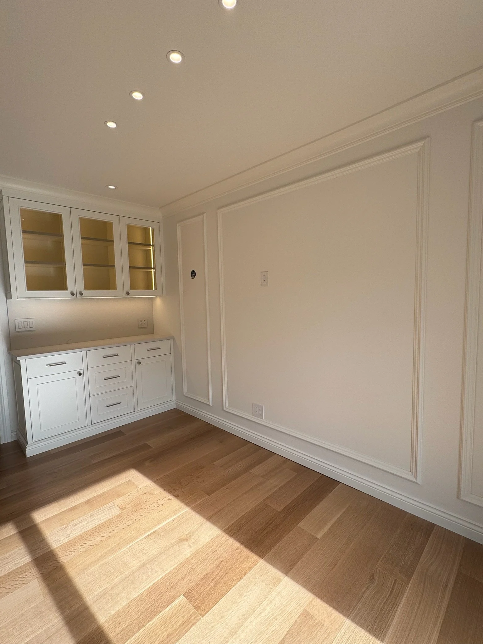 Empty wall with white decorative molding, built-in white cabinets with glass doors, hardwood floor, and ceiling recessed lights.