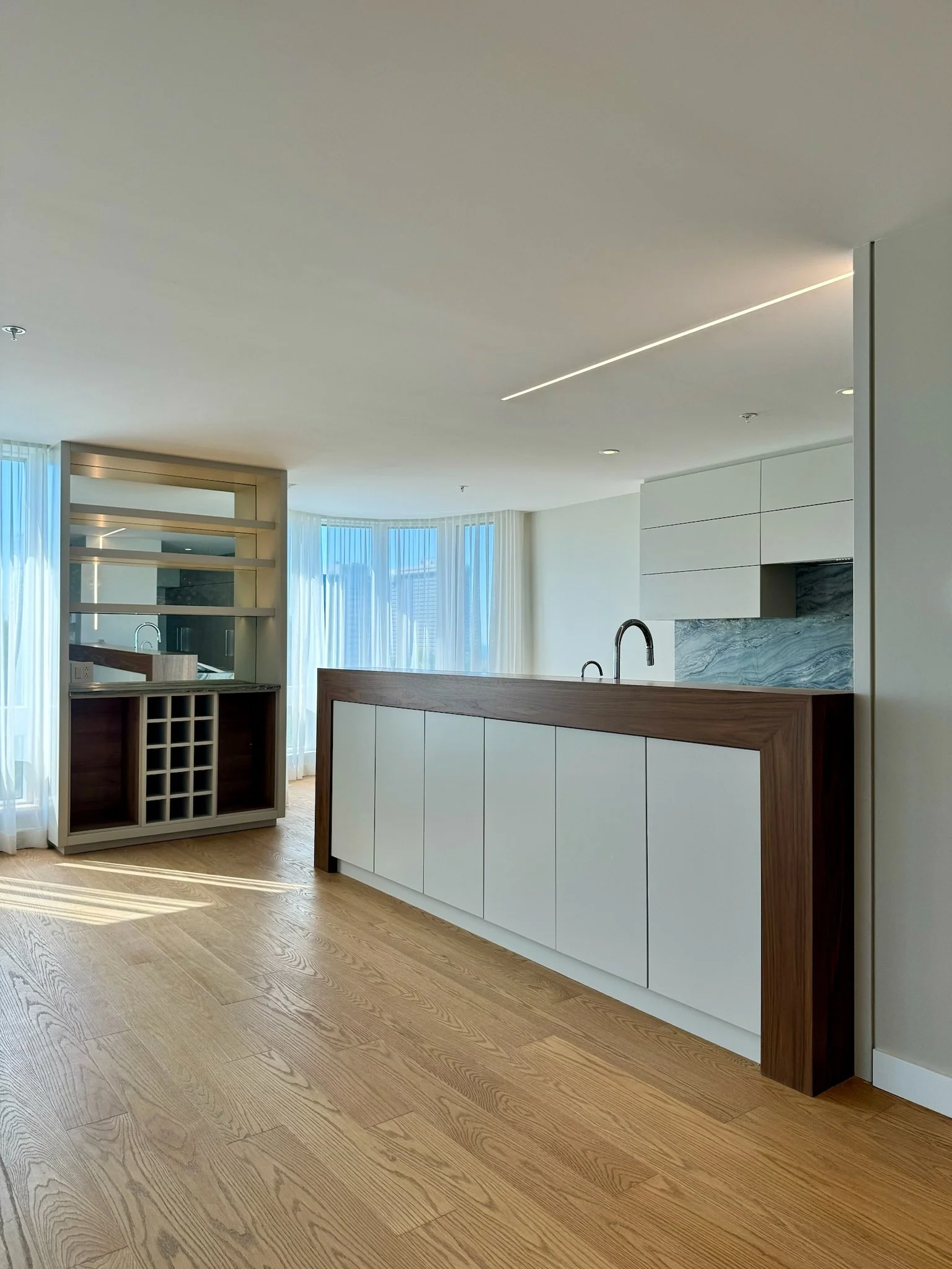 Modern kitchen with a floating island, white cabinets, a marble backsplash, wooden flooring, and floor-to-ceiling windows with sheer white curtains.