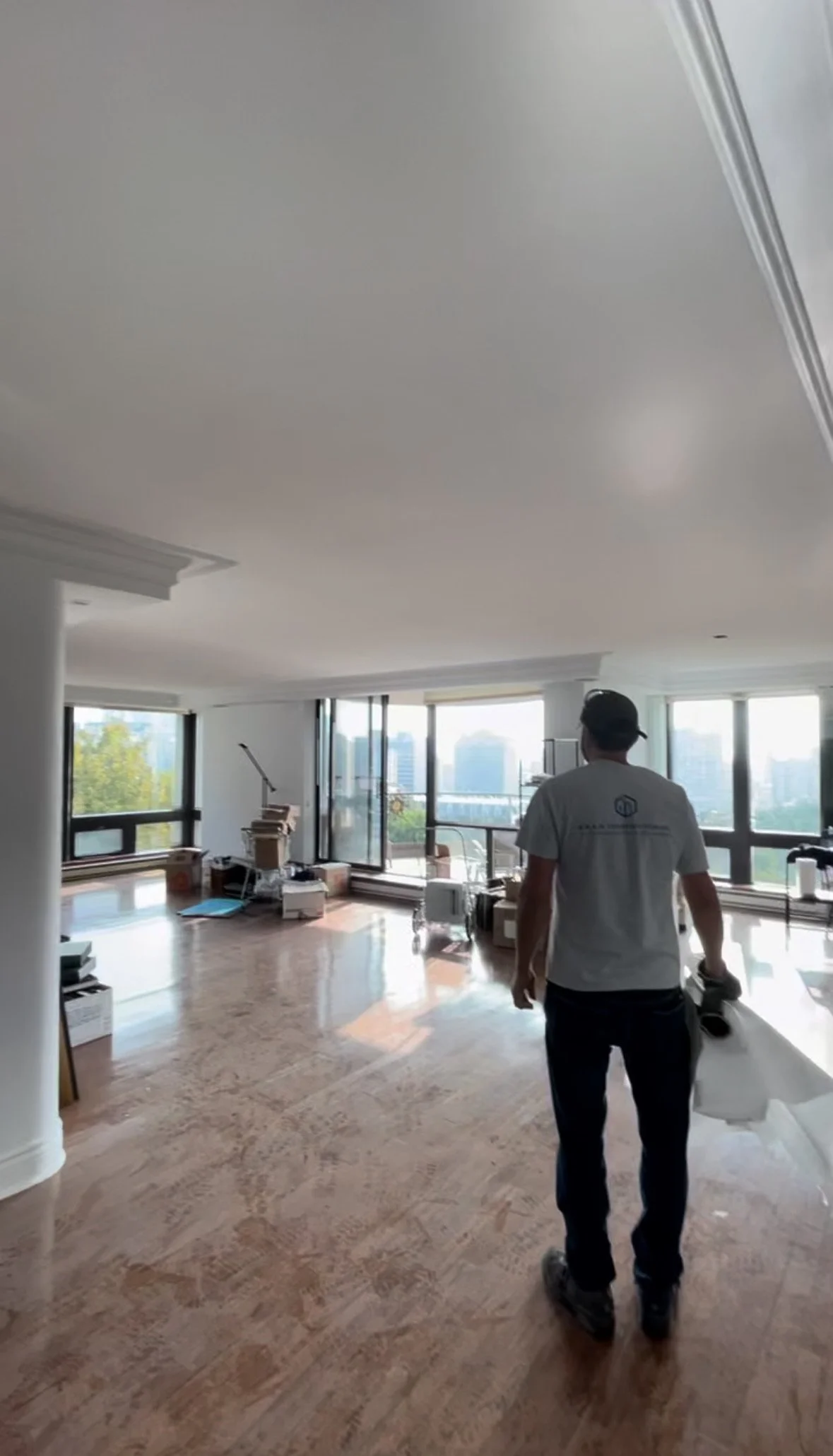 A man walking through an empty, modern apartment or office space with large windows, scattered boxes, and cleaning supplies, during daytime.