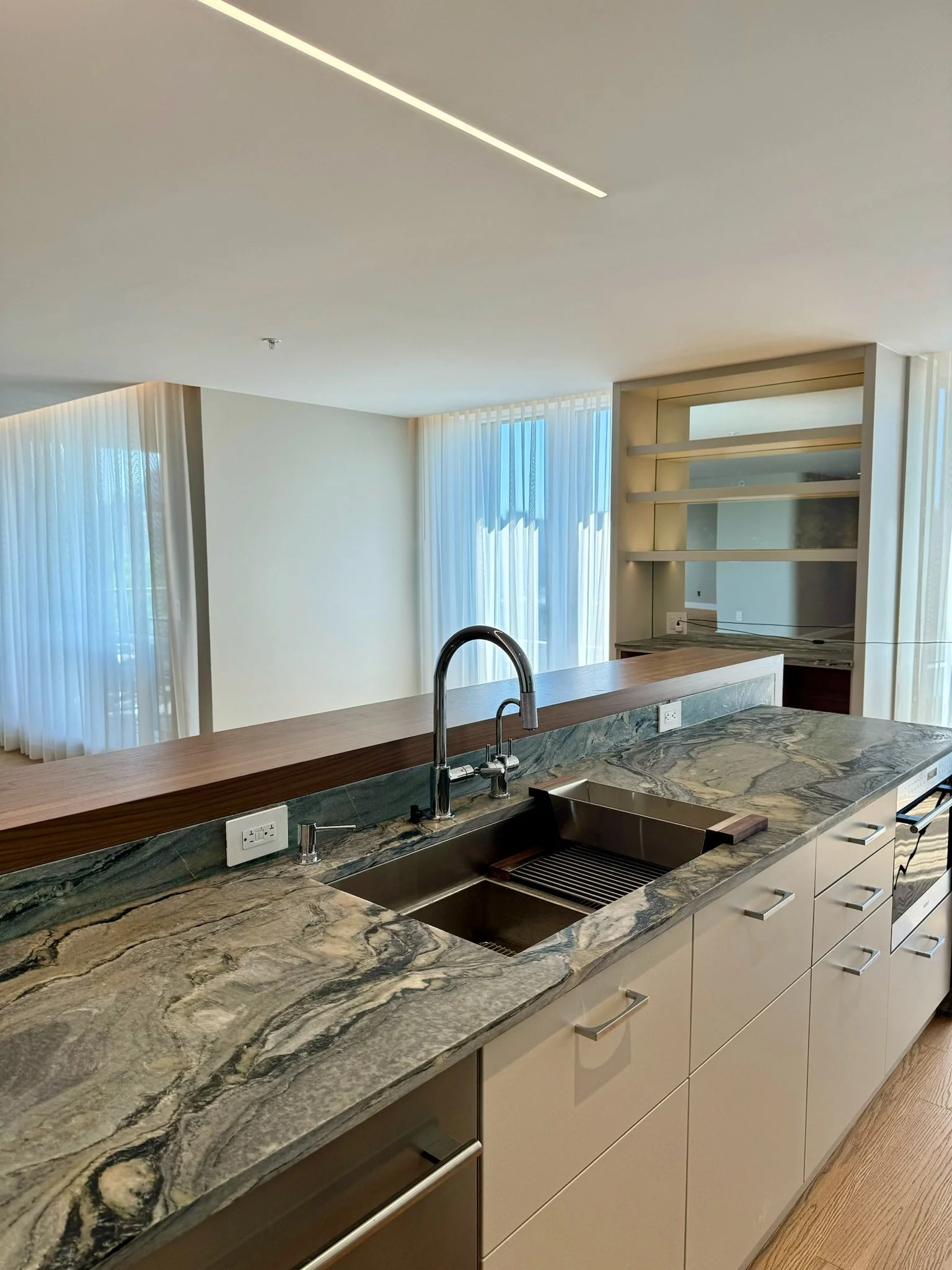 Modern kitchen with a marble countertop, sink, white cabinets, and large windows with sheer curtains.