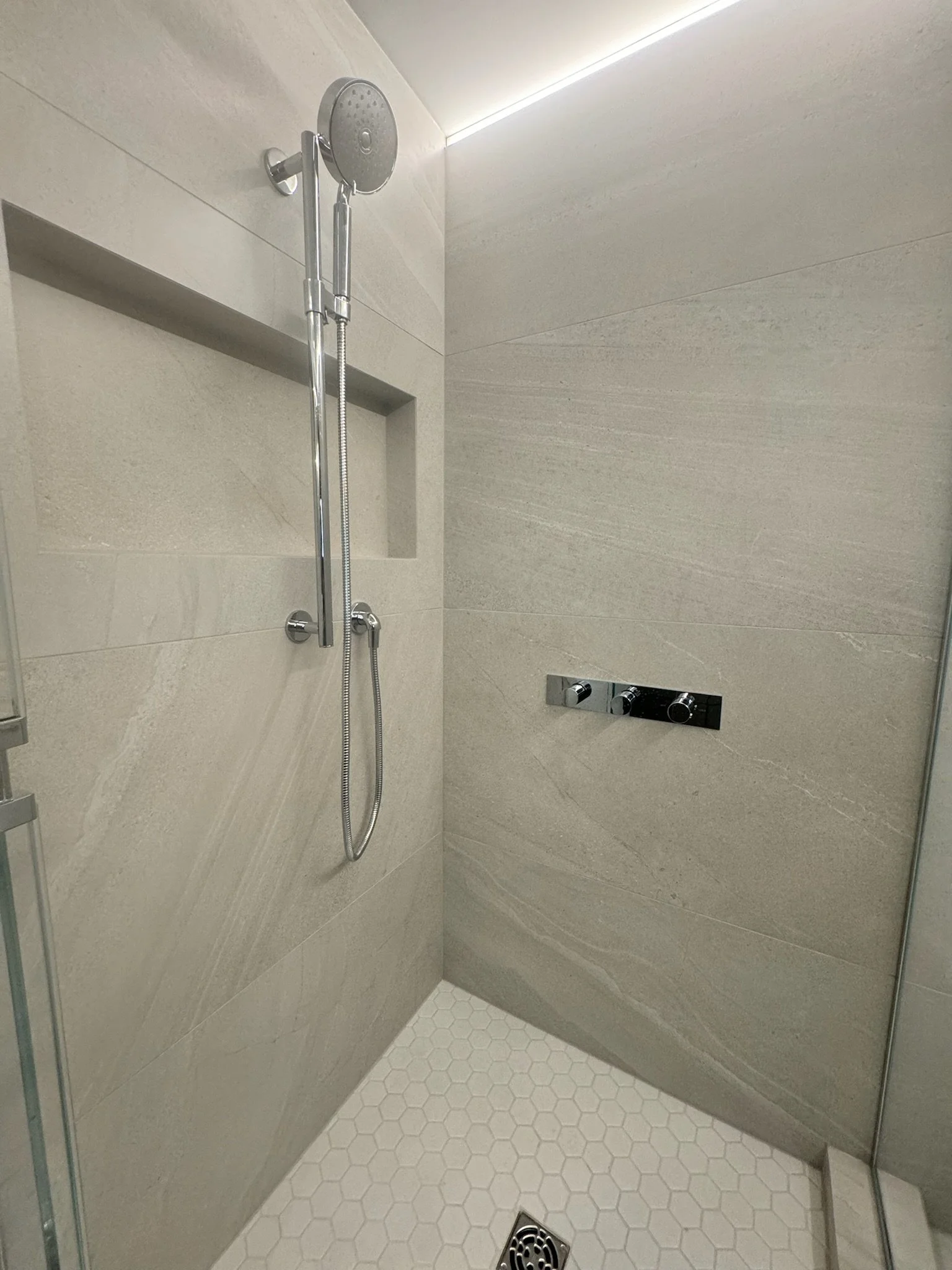 Modern shower with beige tiles, a rainfall shower head, a handheld shower, a recessed shelf, and hexagonal floor tiles.