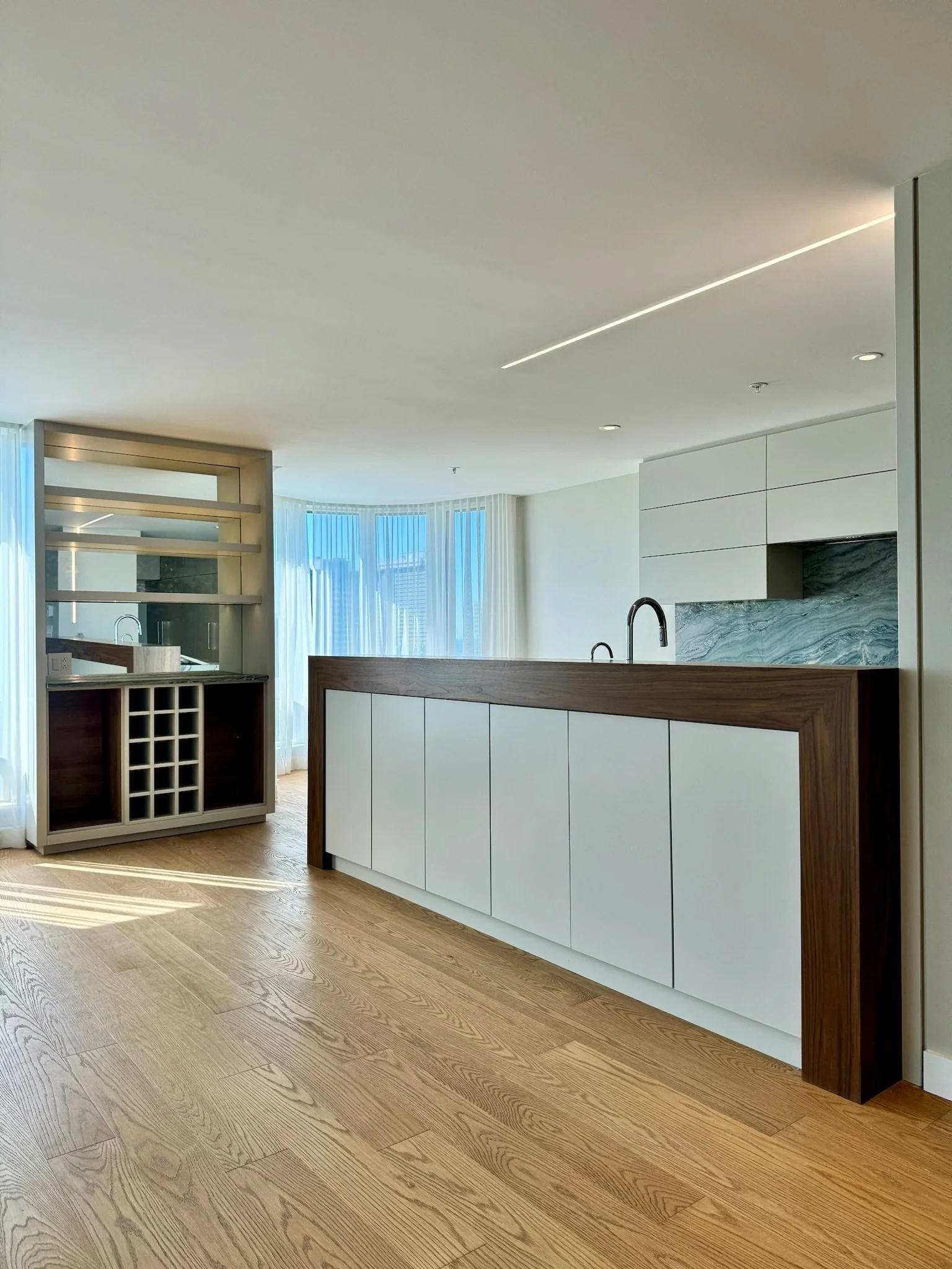 Modern kitchen with white cabinets, marble backsplash, wooden bar, and hardwood floors, with floor-to-ceiling windows and sheer curtains.