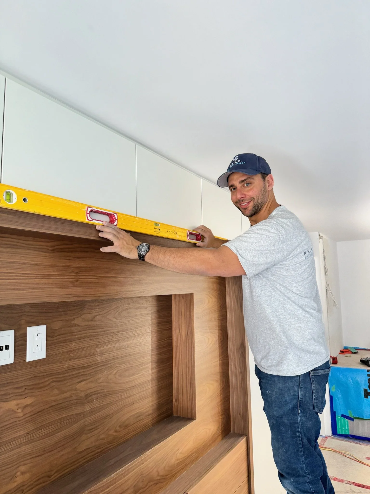 A man in a gray t-shirt, jeans, and a blue cap is installing or adjusting a yellow level on a wooden cabinet.