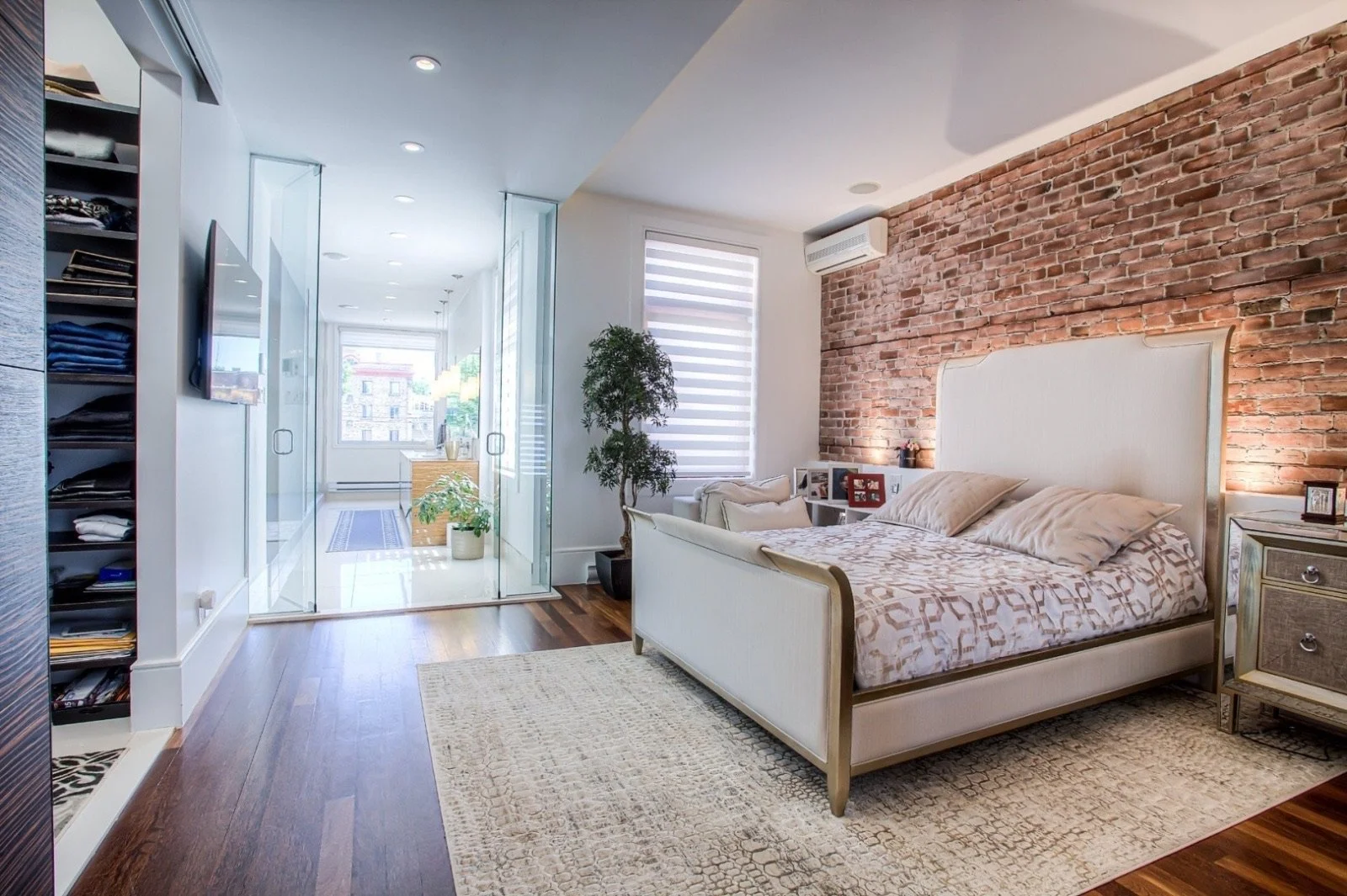 A bedroom with a white bed, brick accent wall, wooden floors, nightstand, window with striped blinds, potted plant, and an office space in the background separated by glass doors.
