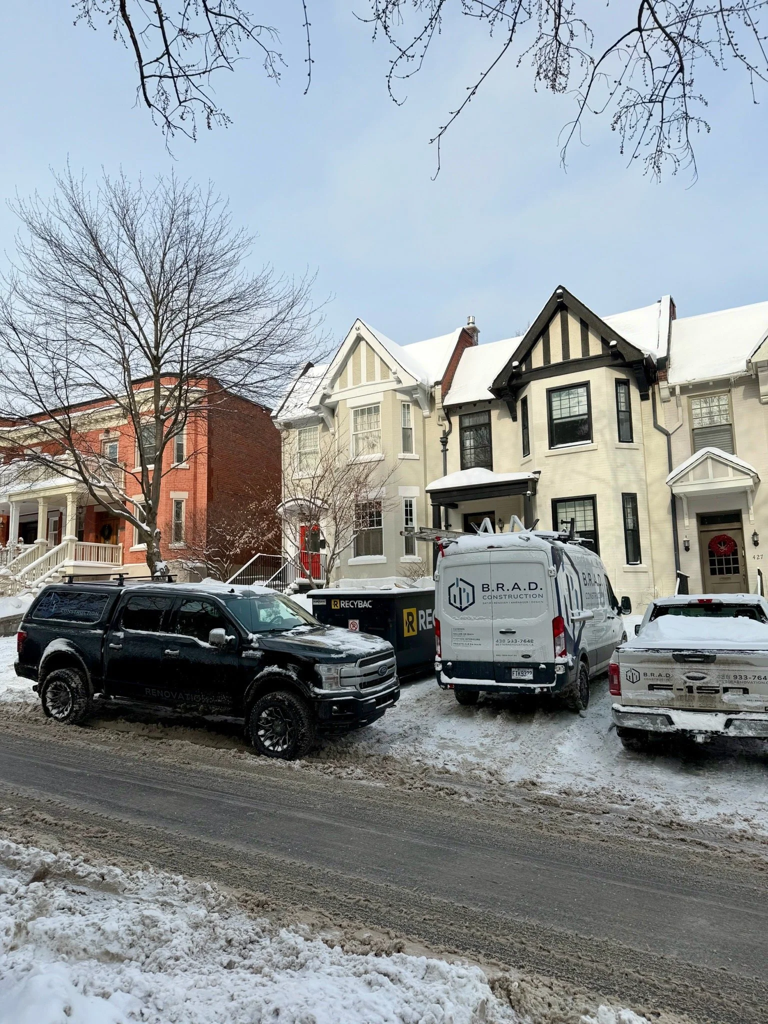 Snow-covered street with parked cars and residential houses, snow on the ground and rooftops, leafless trees, and a partly cloudy sky.