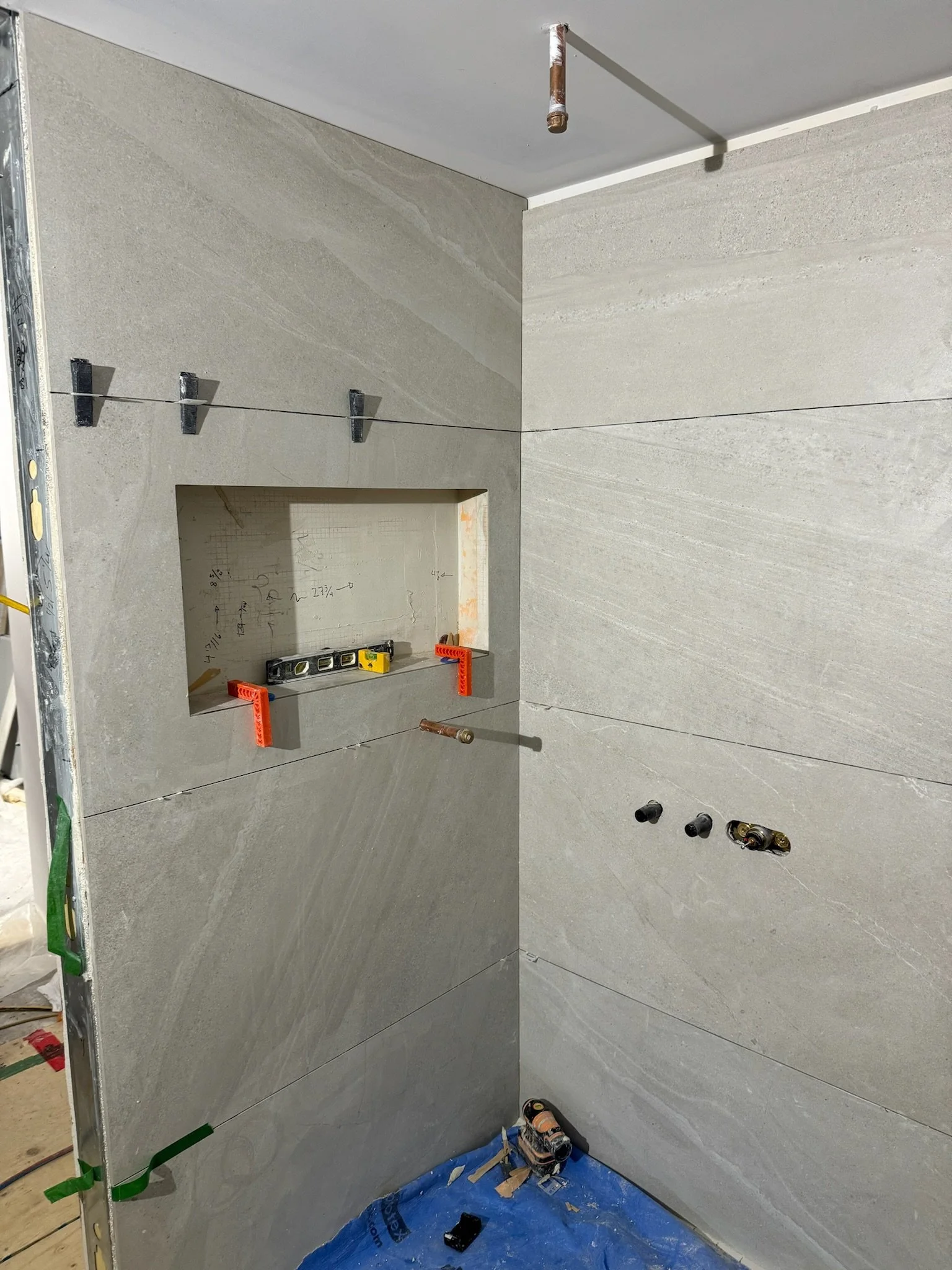 Interior of a bathroom or kitchen under construction, with grey marble-like wall tiles, an electrical outlet, and plumbing pipes. A level tool and orange corner protectors are set on a recessed niche, with a blue plastic sheet on the floor.