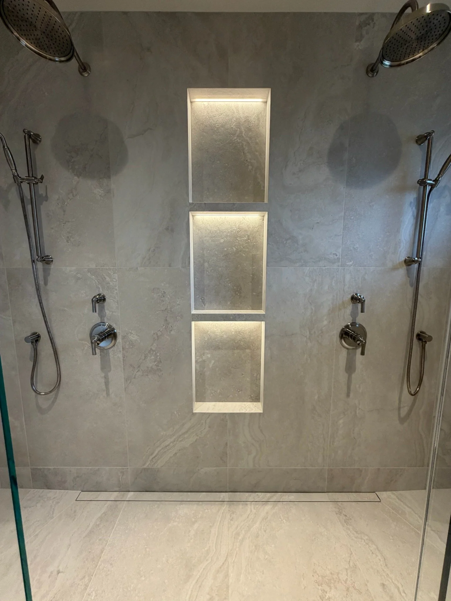 A modern shower area with two shower heads on either side, and three built-in shelves with lighting in the center wall. The shower has beige stone tiles on the walls and floor.