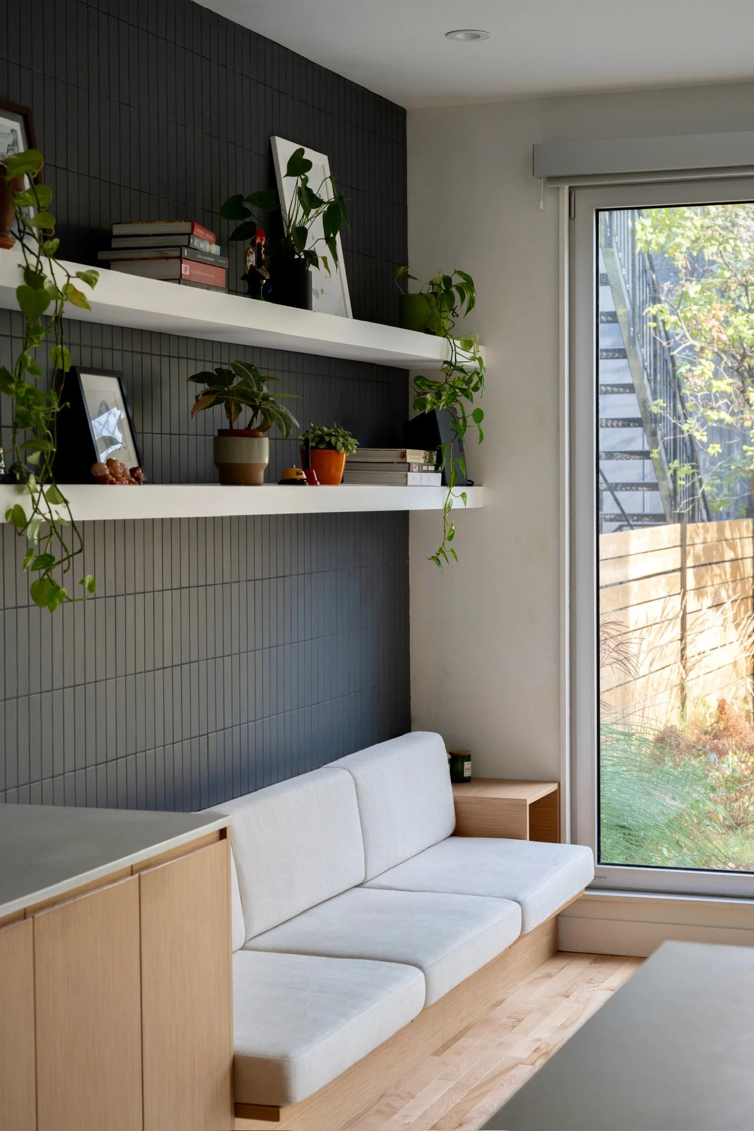 Modern living room with white sofa, light wood furniture, gray tile wall with black accents, potted plants on white floating shelves, large window with city view, outside greenery.
