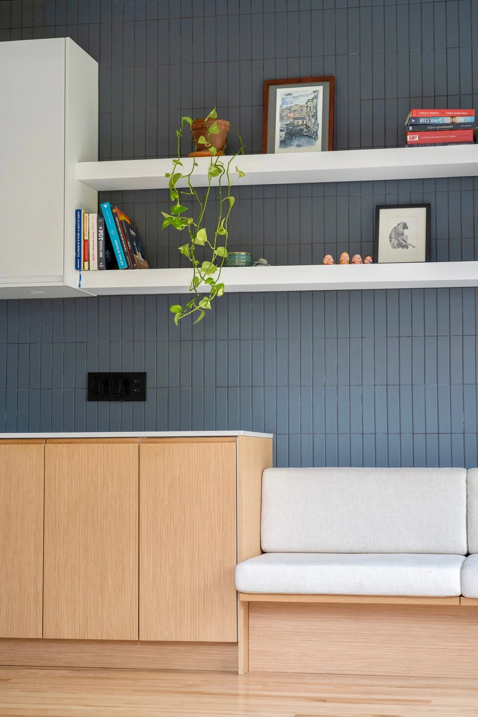 Interior scene with blue tiled wall, white shelving, framed artwork, a potted plant with trailing vines, a small animal figurine, and a light-colored sofa.