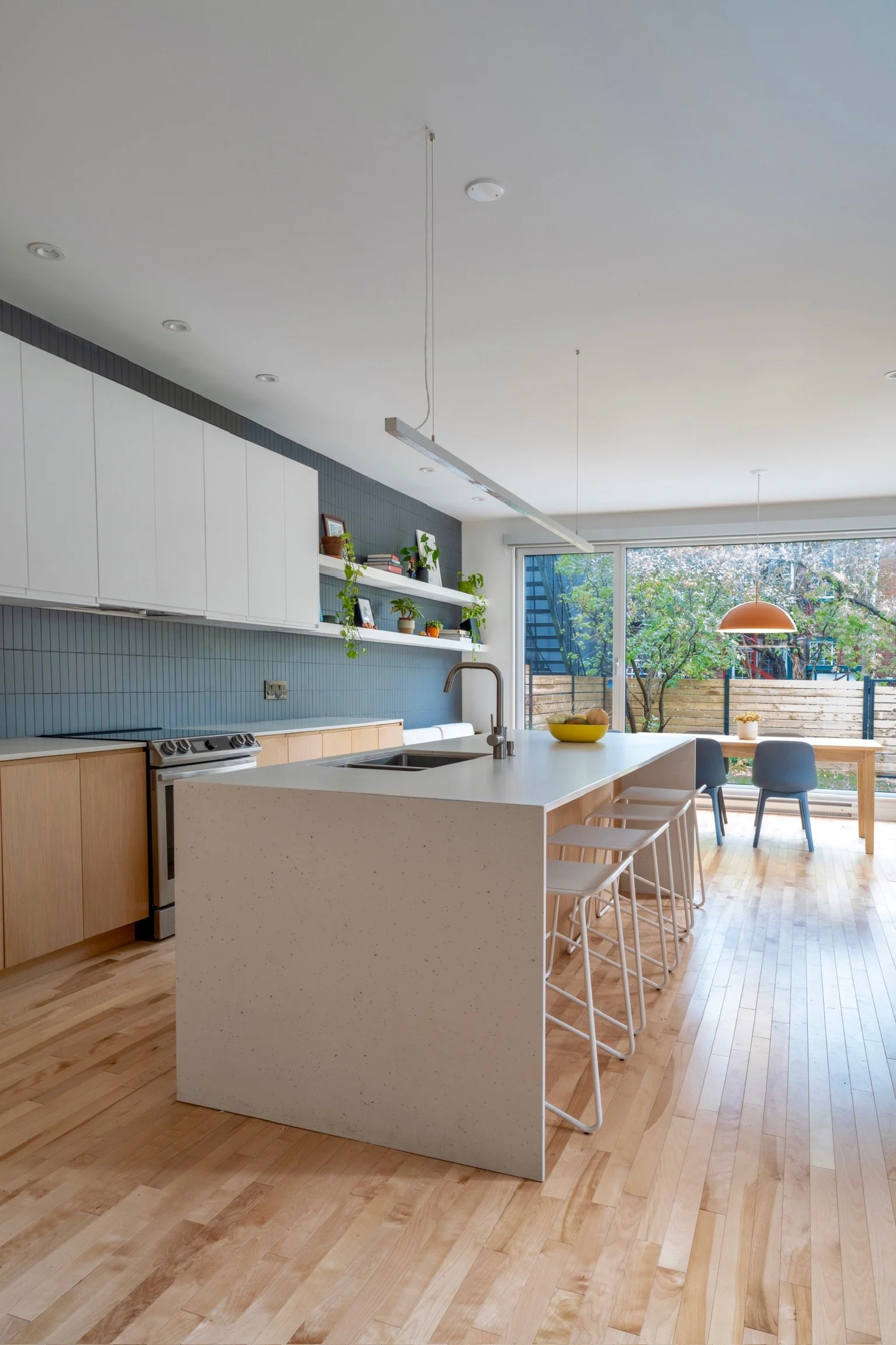 Modern kitchen with white cabinetry, a large island with seating, wood flooring, and a dining area near large windows with a view of greenery outside.