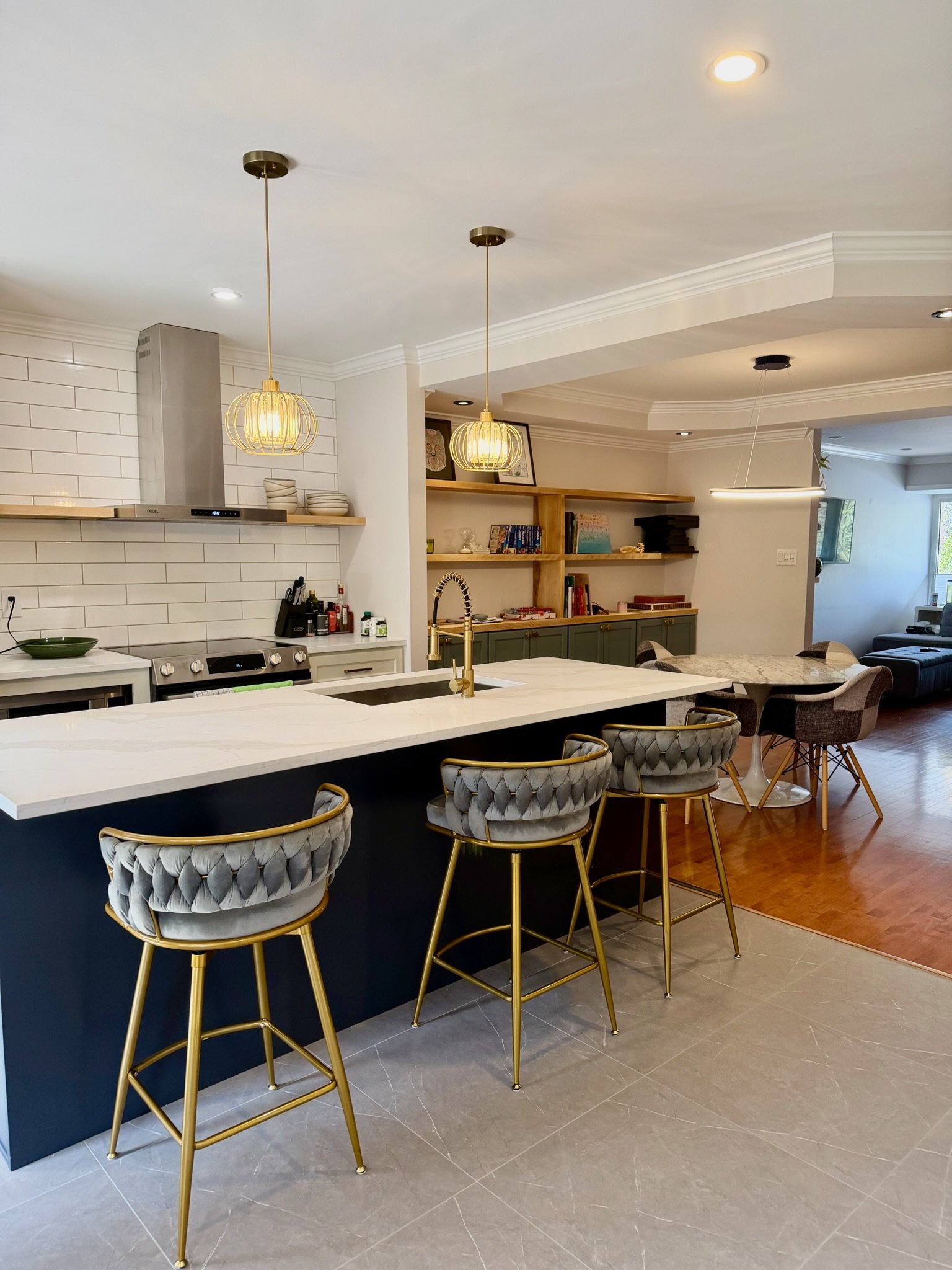 Modern kitchen with white island, gold and gray chairs, open shelving with books and decor, white tiled wall, and partial view of living area with large window.