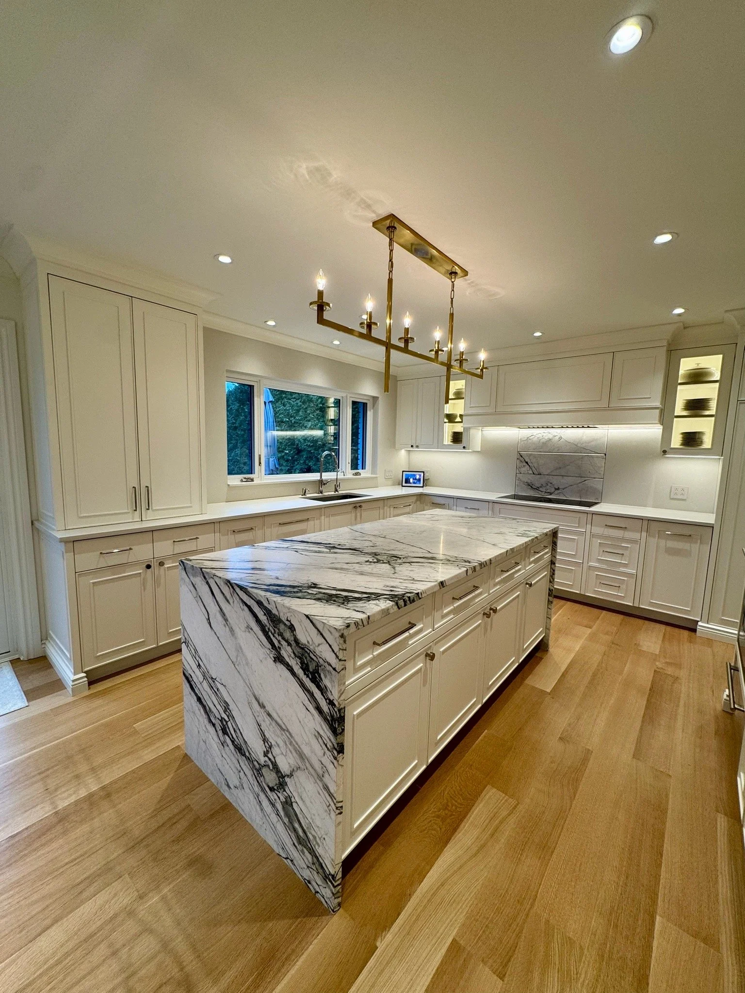 Modern white kitchen with marble island, white cabinets, pendant chandelier, and wooden floor.