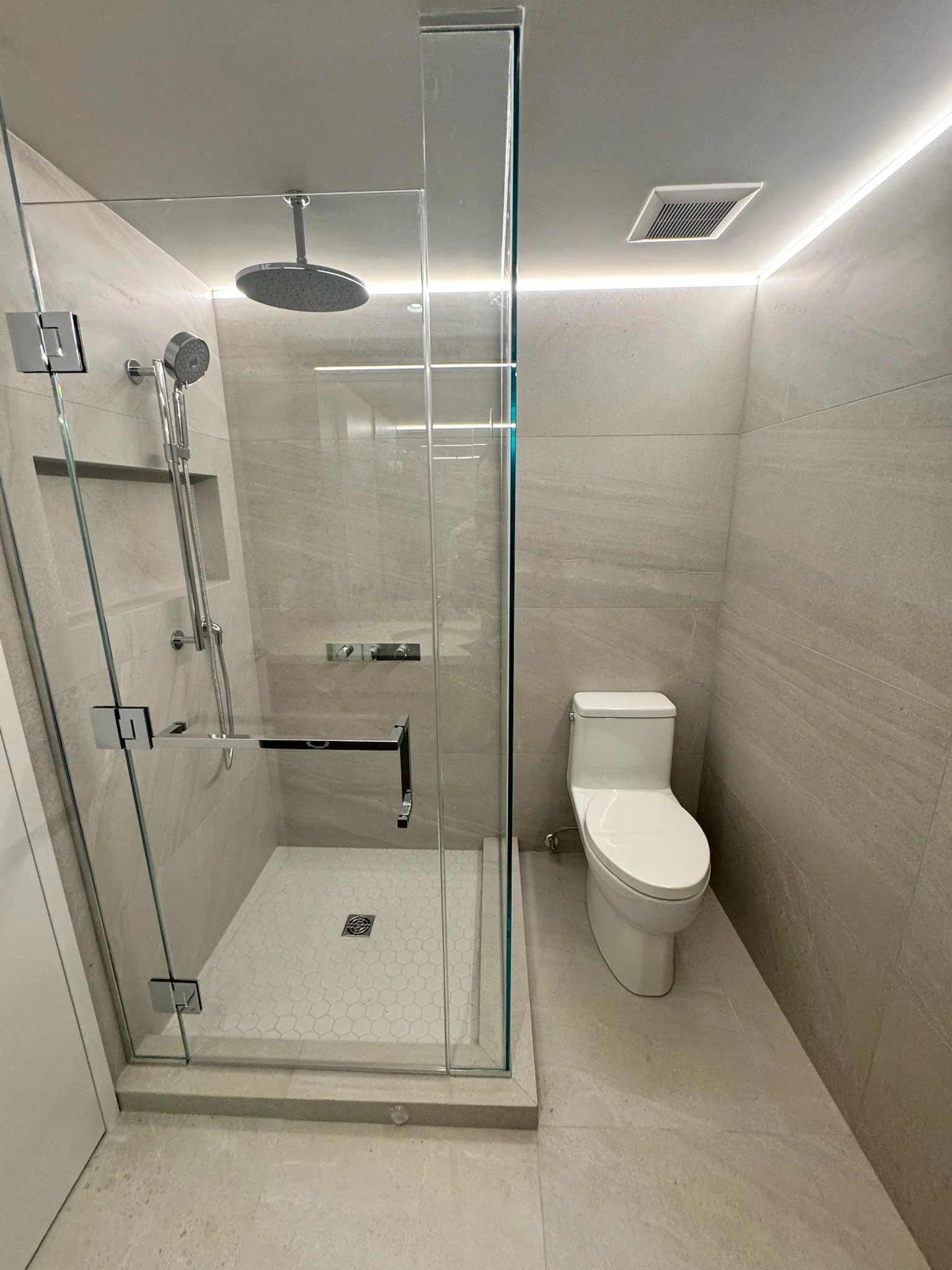 Modern bathroom with glass-enclosed shower, rainfall showerhead, handheld shower, white toilet, beige tiled walls, and minimal decor.