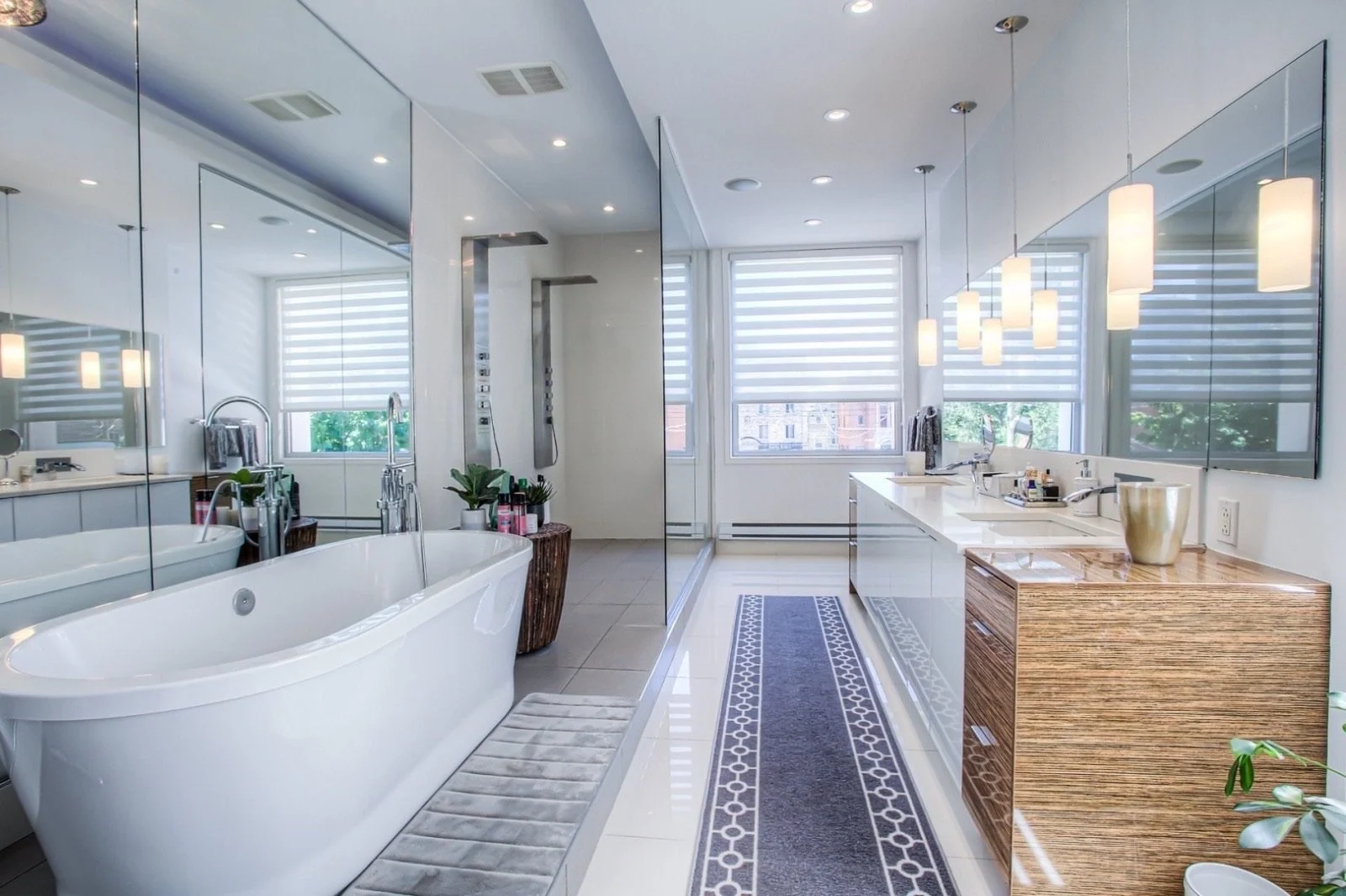 Modern bathroom with a white bathtub, a glass shower, large windows with blinds, and a double sink vanity with pendant lights.