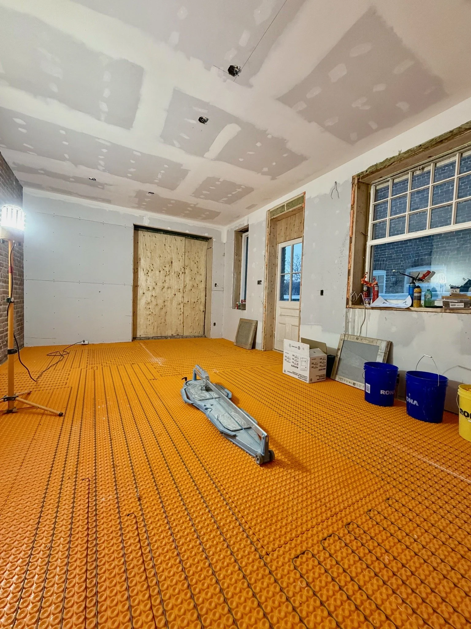 A room under renovation with drywall ceiling and walls, a window, and a sliding door. The floor is covered with orange anti-slip heating cable mats. Tools, buckets, and boxes are scattered around.