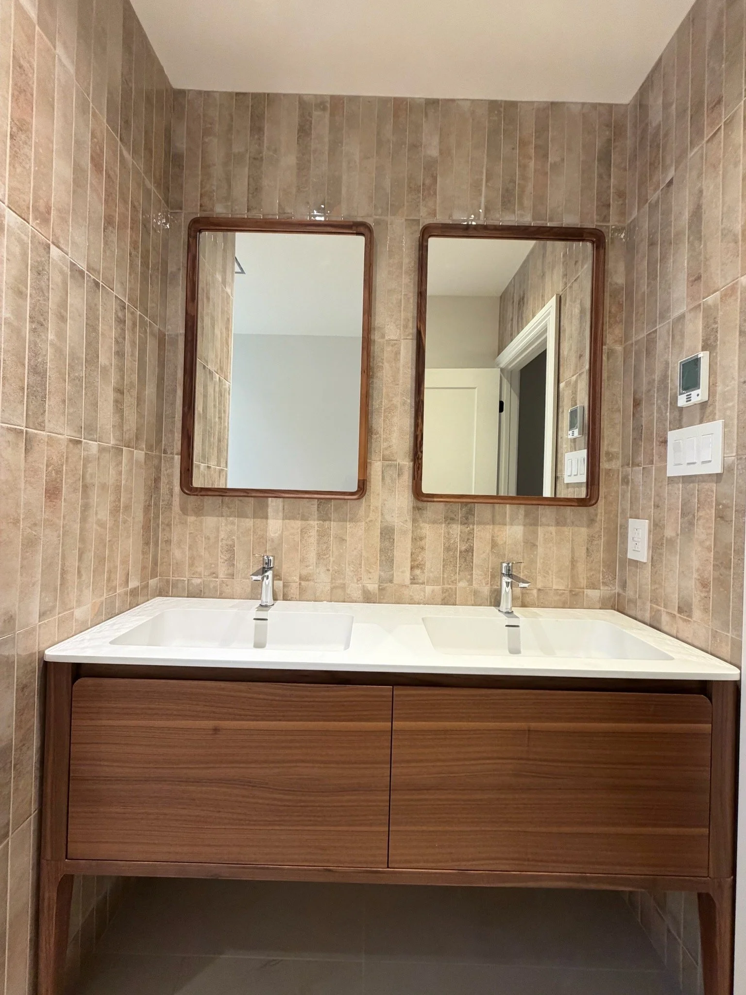 A modern double bathroom vanity with a white countertop and two rectangular sinks, a wood cabinet, and two mirrored wall cabinets, set against beige tiled walls.