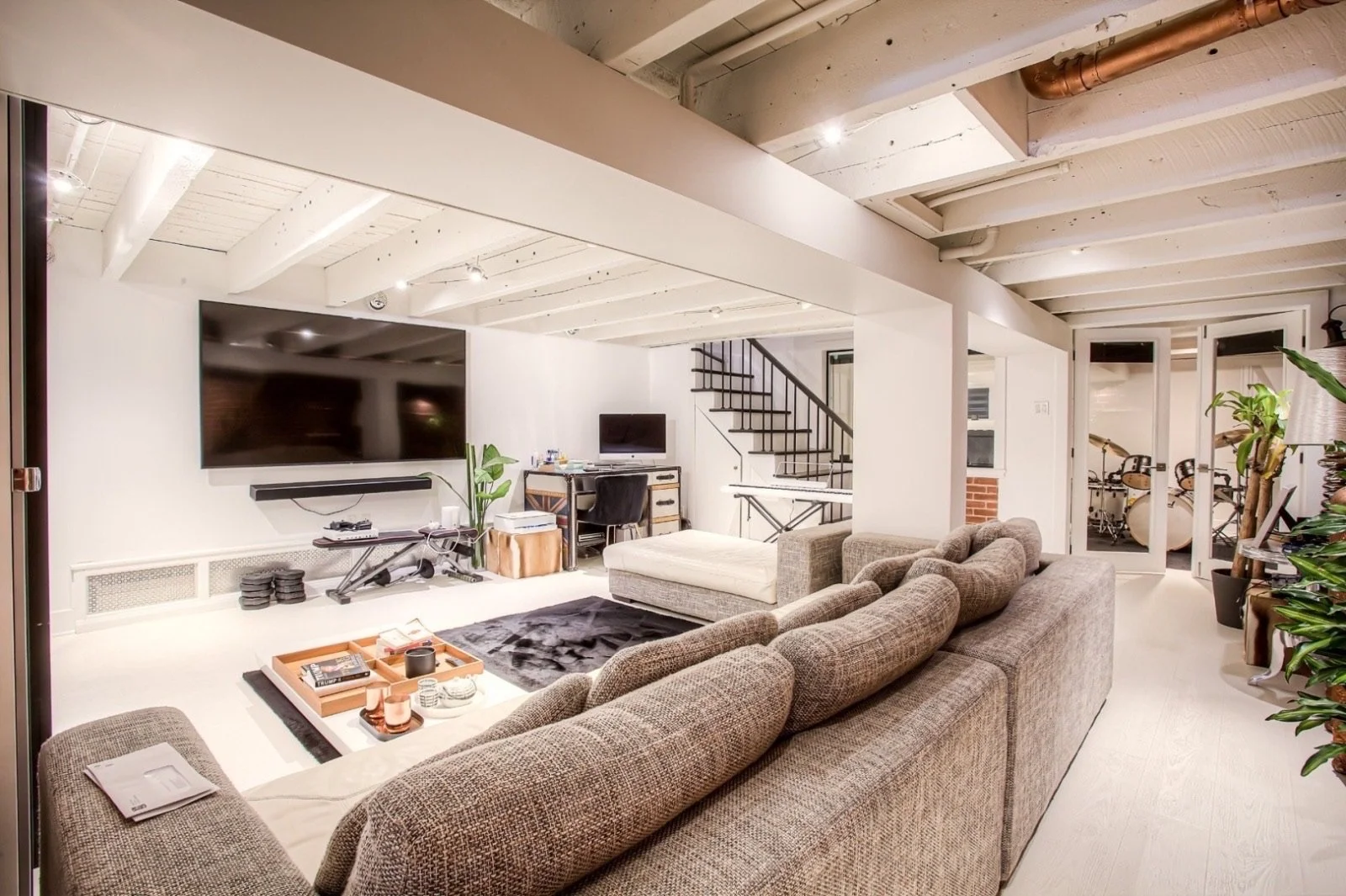 A cozy living room with a large brown sectional sofa facing a flat-screen TV mounted on the wall. The room has white walls and ceiling with exposed beams, a staircase with black railing, indoor plants, and a door leading to a room with drums and an e