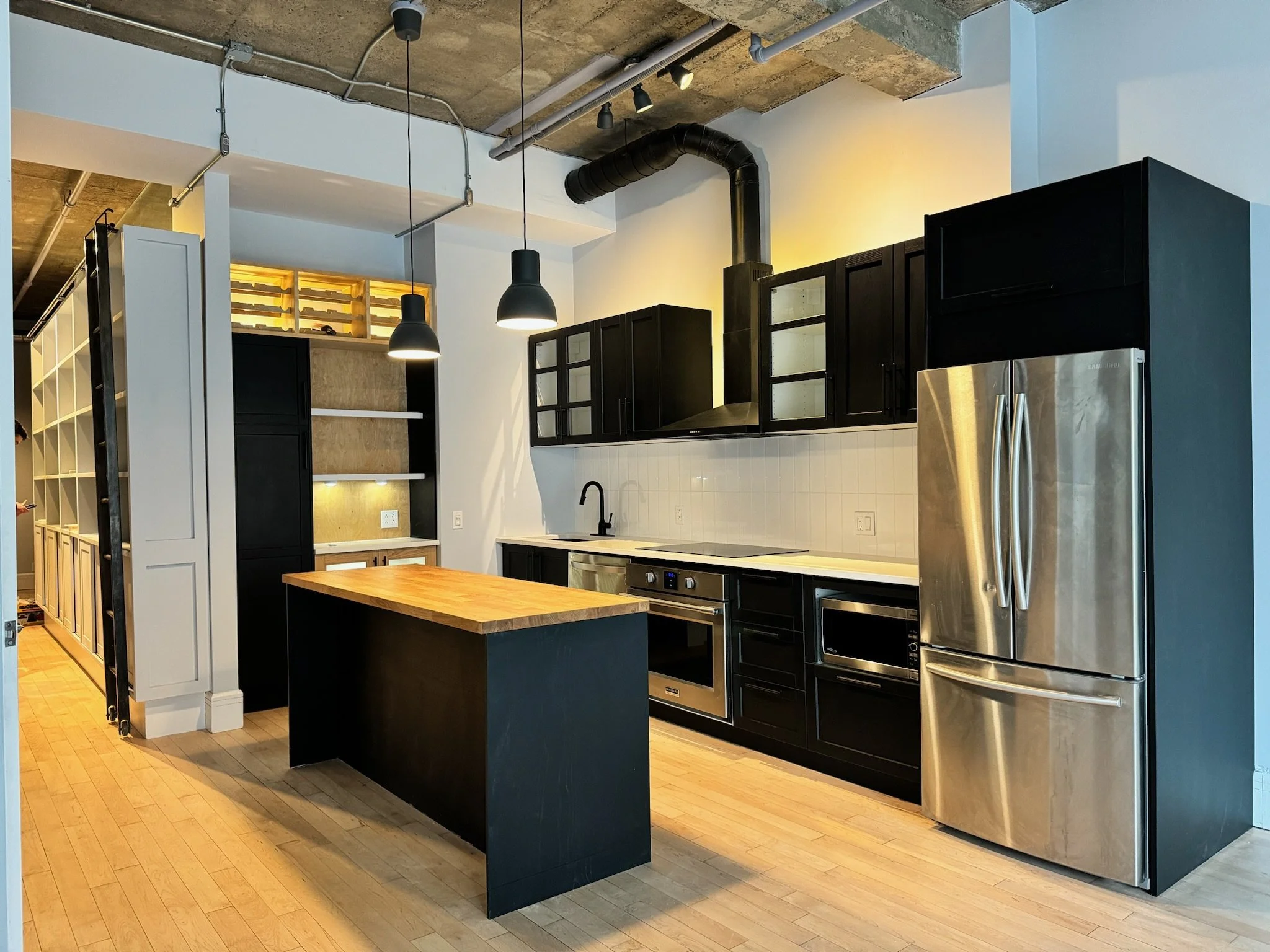 Modern kitchen with black cabinets, stainless steel refrigerator, wooden kitchen island, pendant lighting, and exposed concrete ceiling.