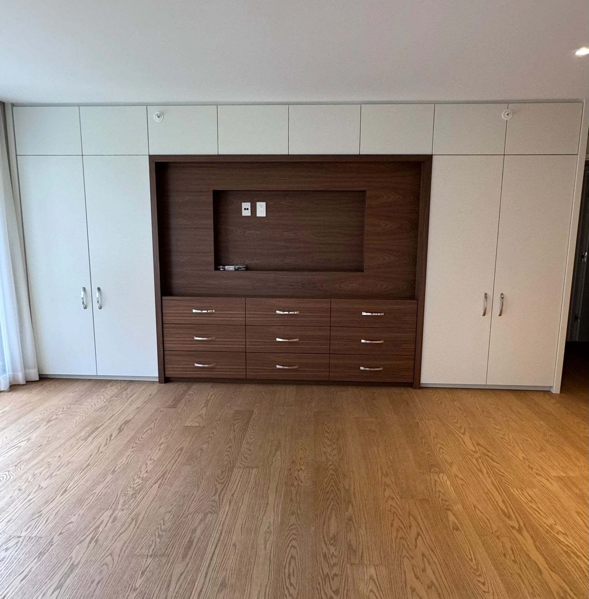 A modern room with wood flooring, a wall with white cabinets, and a wooden panel with electrical outlets and a TV mount.