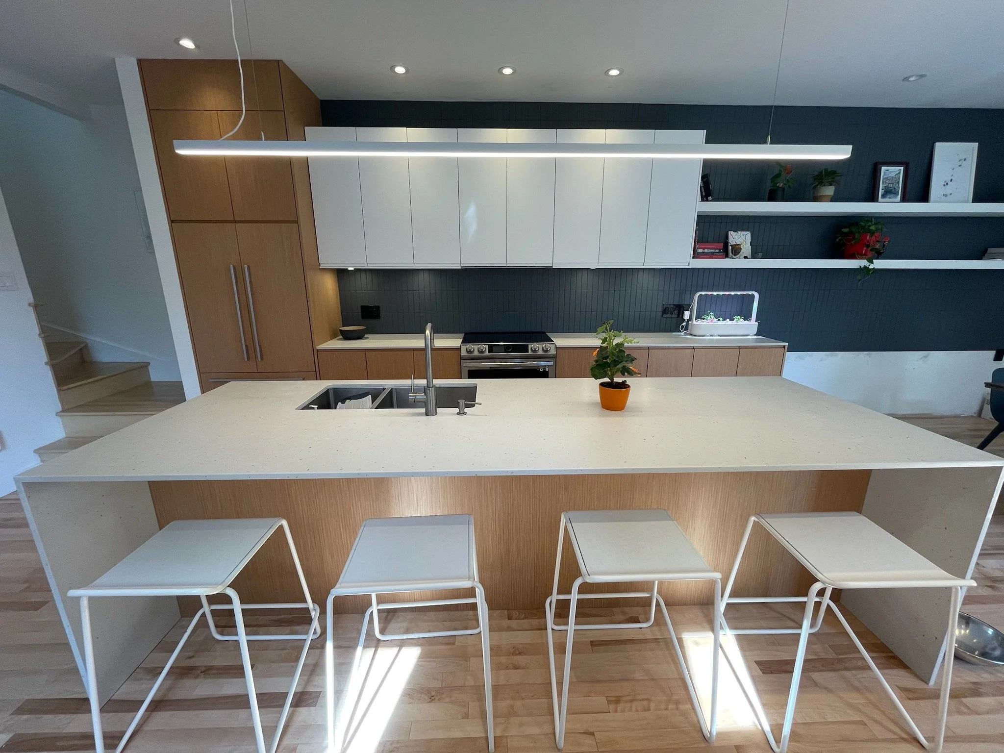 Modern kitchen with a large white island, wooden cabinets, floating white shelves, dark backsplash, potted plants, artwork, and track lighting.