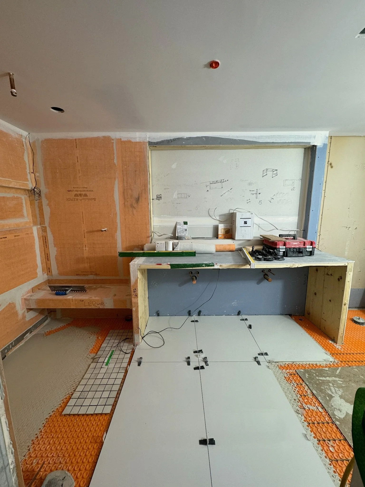 Indoor construction site showing partially tiled floor, exposed framing, and unfinished walls. Tools and materials are scattered around.