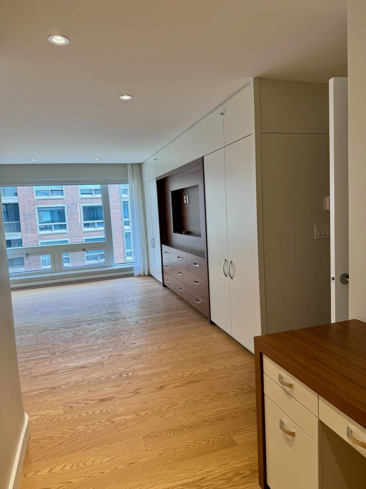 Interior of a modern apartment with large window, wooden floors, built-in cabinets, and a television mounted on the wall.