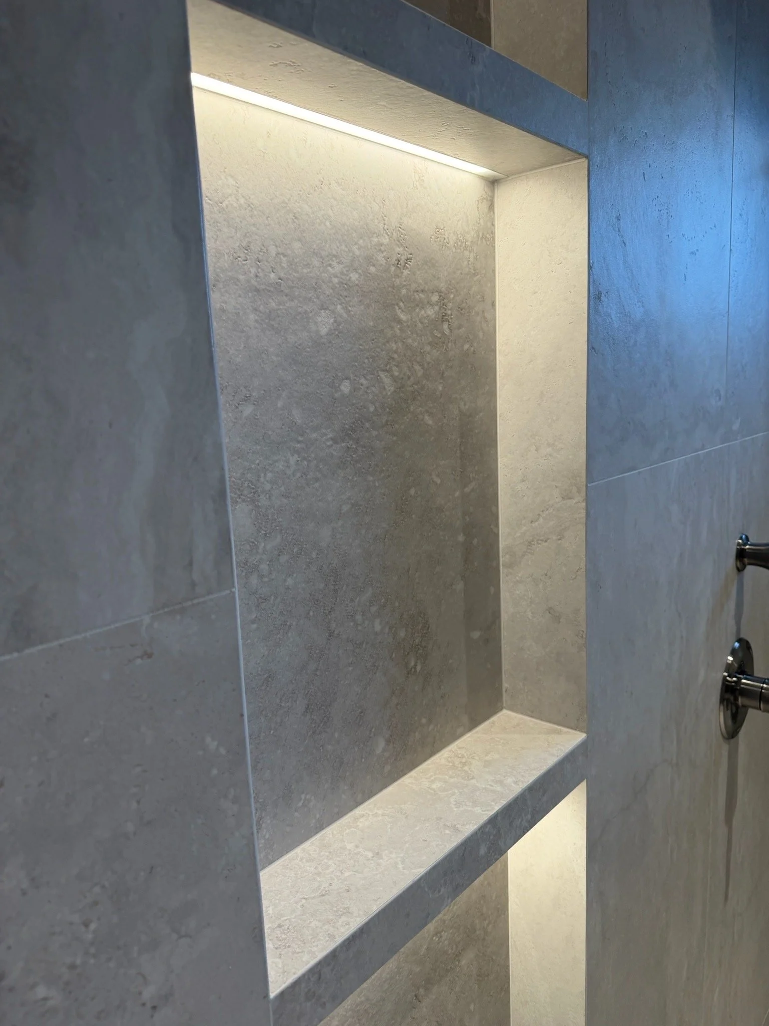 A modern bathroom with a shower area featuring grey textured tile walls and a built-in shelf. The shower has a minimalistic design with a simple chrome handle visible on the right side.