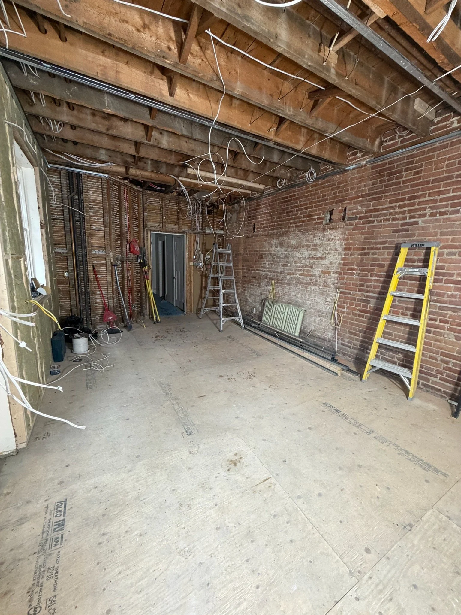 Interior of a room under construction with exposed brick walls, unfinished ceiling with wooden beams and electrical wiring, and a concrete floor. Construction tools and materials are present, including a yellow ladder and a workbench.