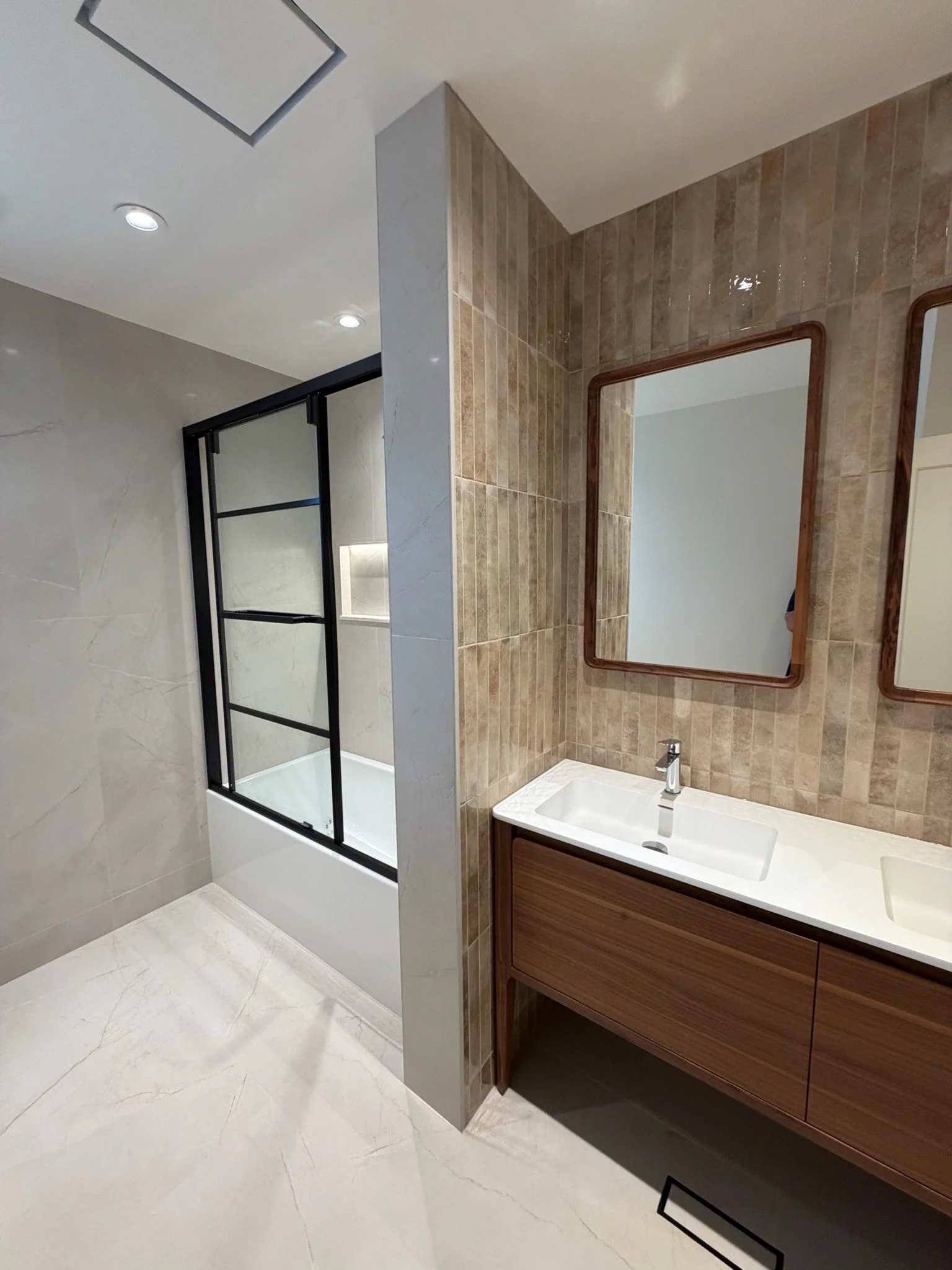 A modern bathroom with a shower area on the left with a glass enclosure and black frame, beige tiled walls, a white sink with a wooden vanity, and two rectangular mirrors above the sink. The floor has large light-colored tiles.