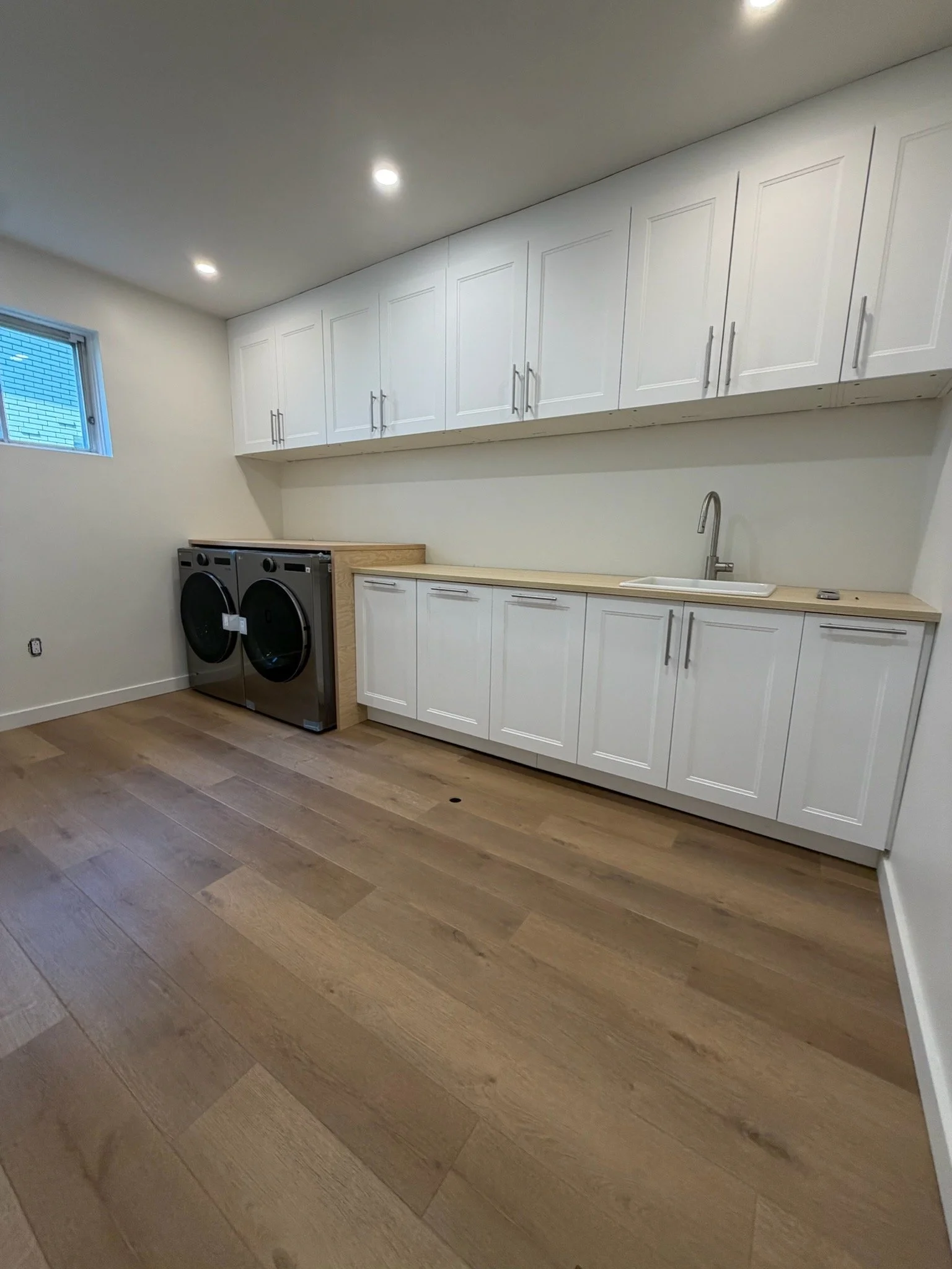 Laundry room with white cabinets, a sink, a washer and dryer, and a small window, with wood flooring and recessed ceiling lights.
