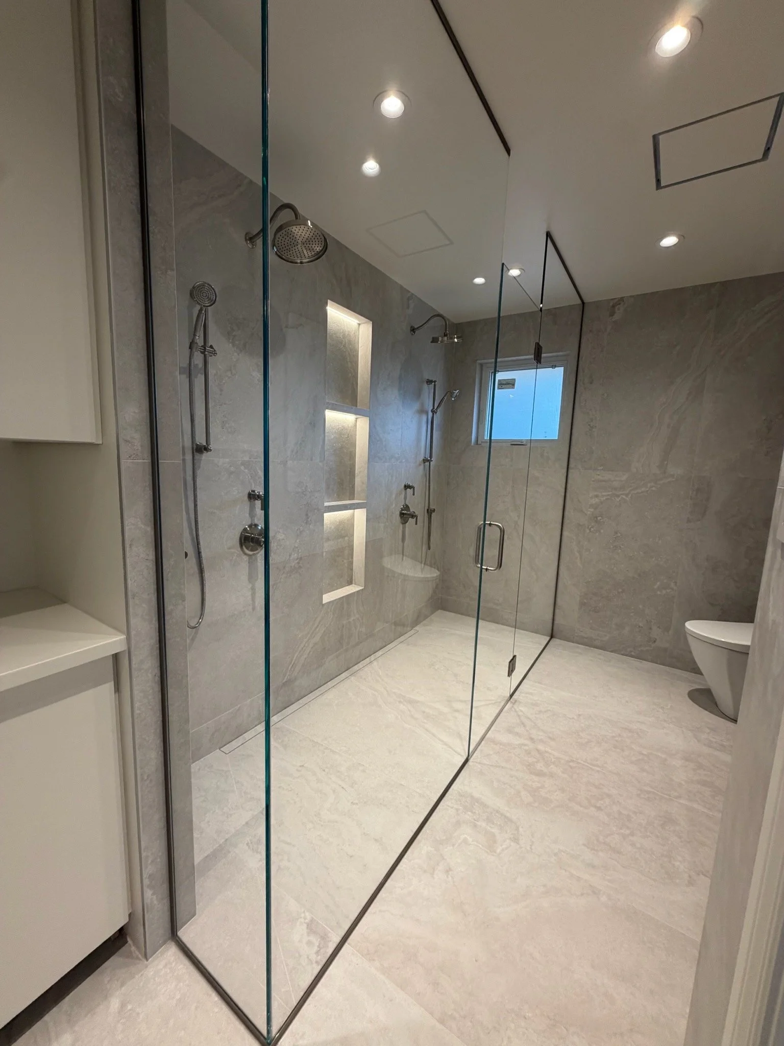 Modern bathroom with a walk-in shower enclosed by glass panels, featuring multiple showers including a rain showerhead and handheld shower, gray marble walls with built-in shelves, a small window, beige floor tiles, and a modern toilet.