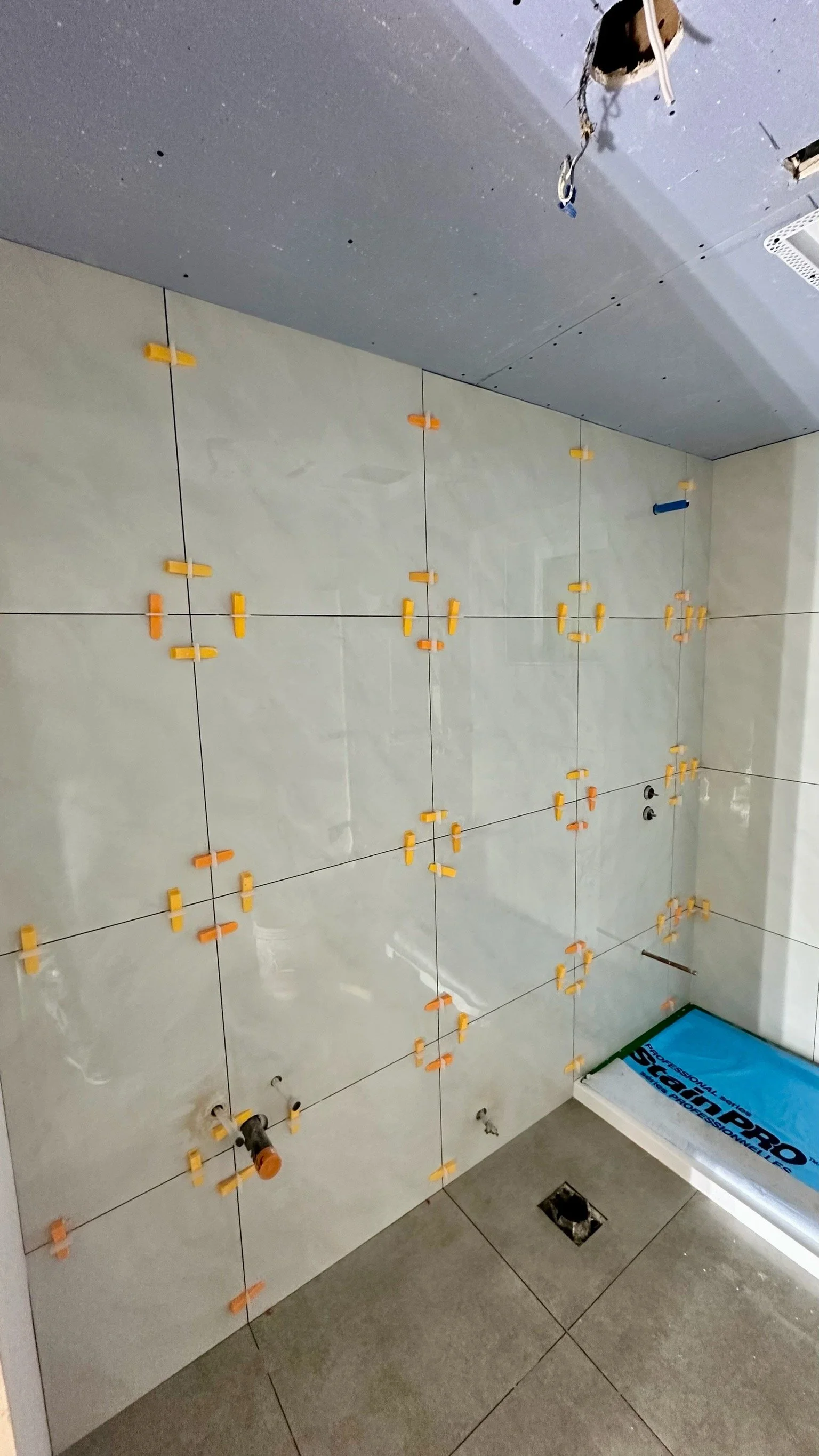 Bathroom wall with partially installed tiles and yellow spacers, with plumbing fixtures and electrical wires visible.